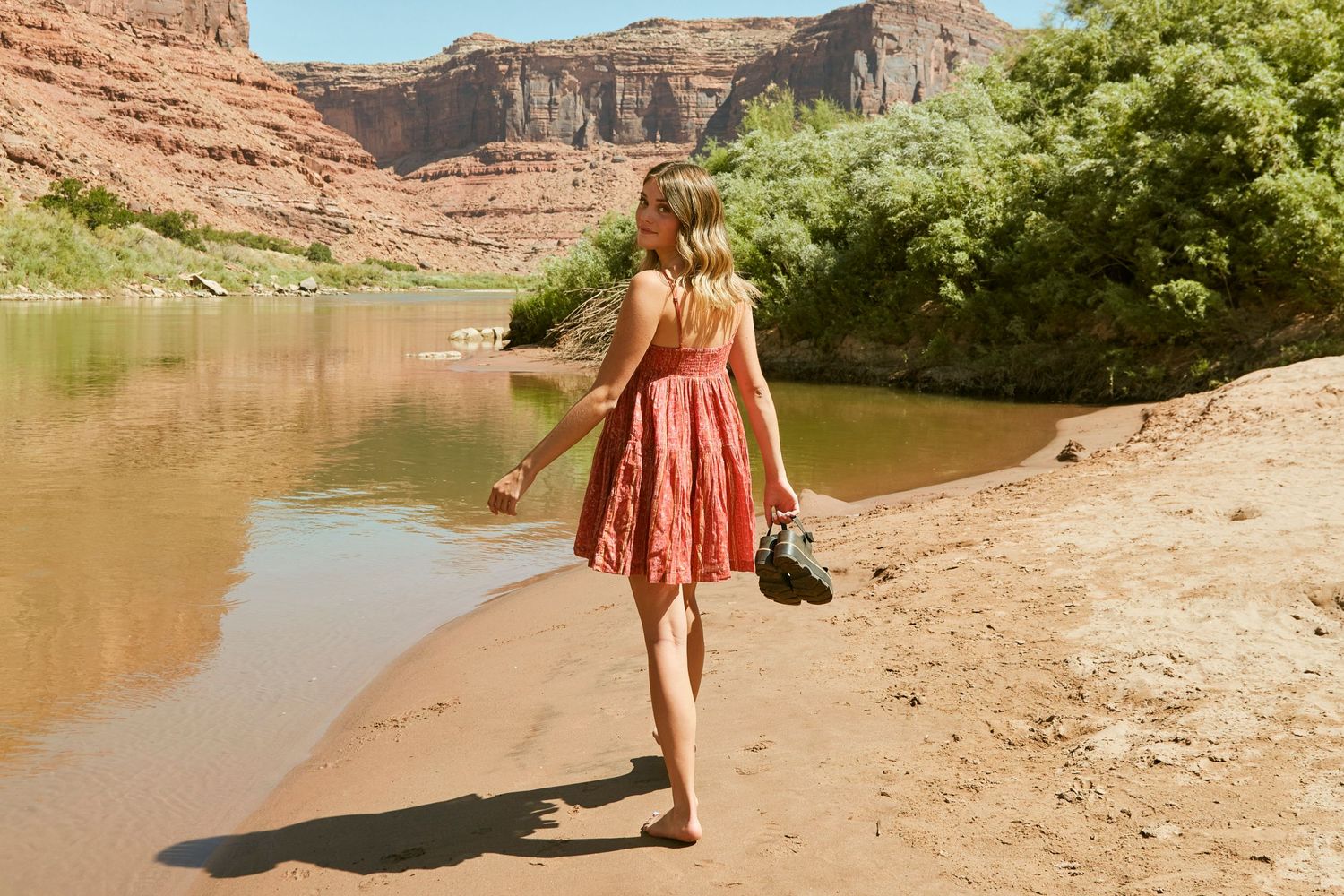 A red sundress flows in the breeze along a scenic riverbank with red rock cliffs.