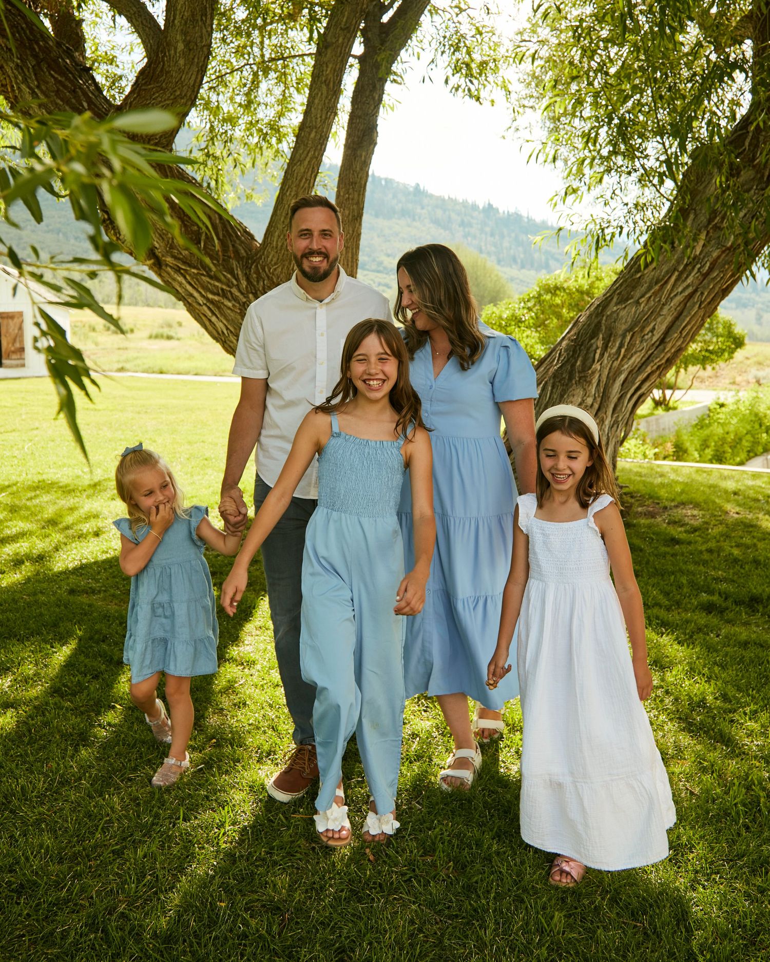 A family wearing coordinated blue and white outfits poses together under a large tree in summer.