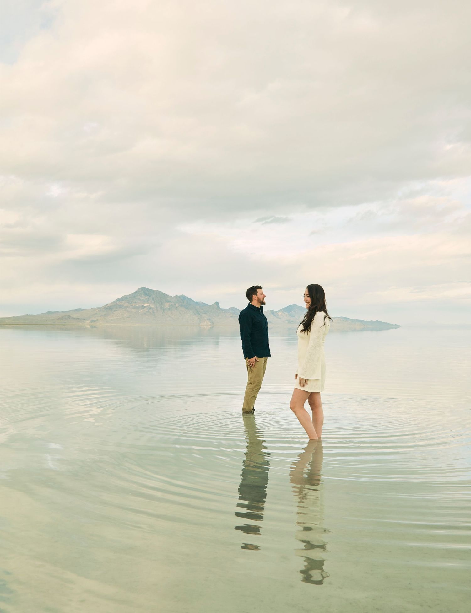 Couple standing in shallow water with mountain landscape in soft, dreamy lighting.