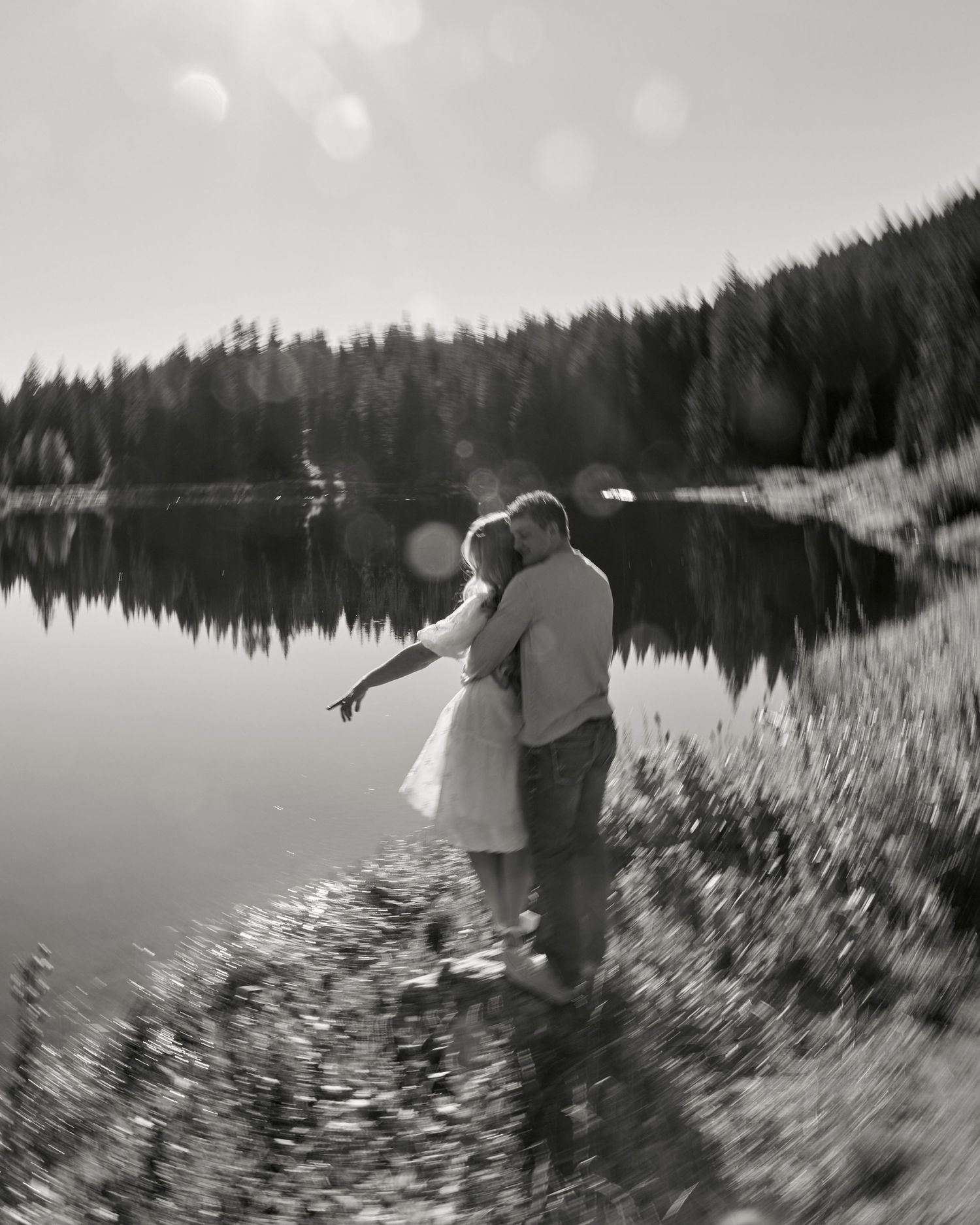 Black and white image of couple standing by misty lake with forest reflection.