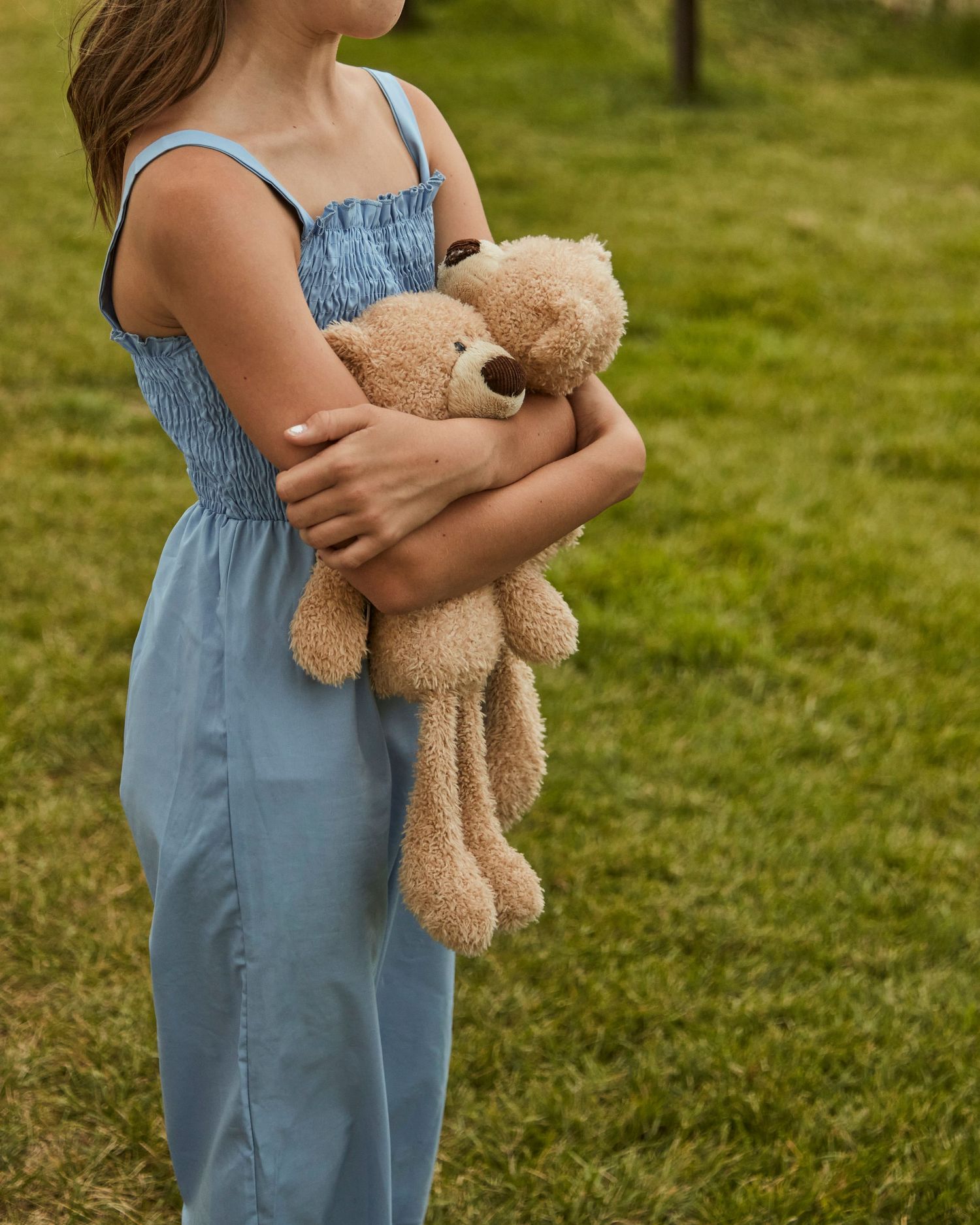 Person in blue denim overalls holding a soft brown teddy bear in a grassy field.