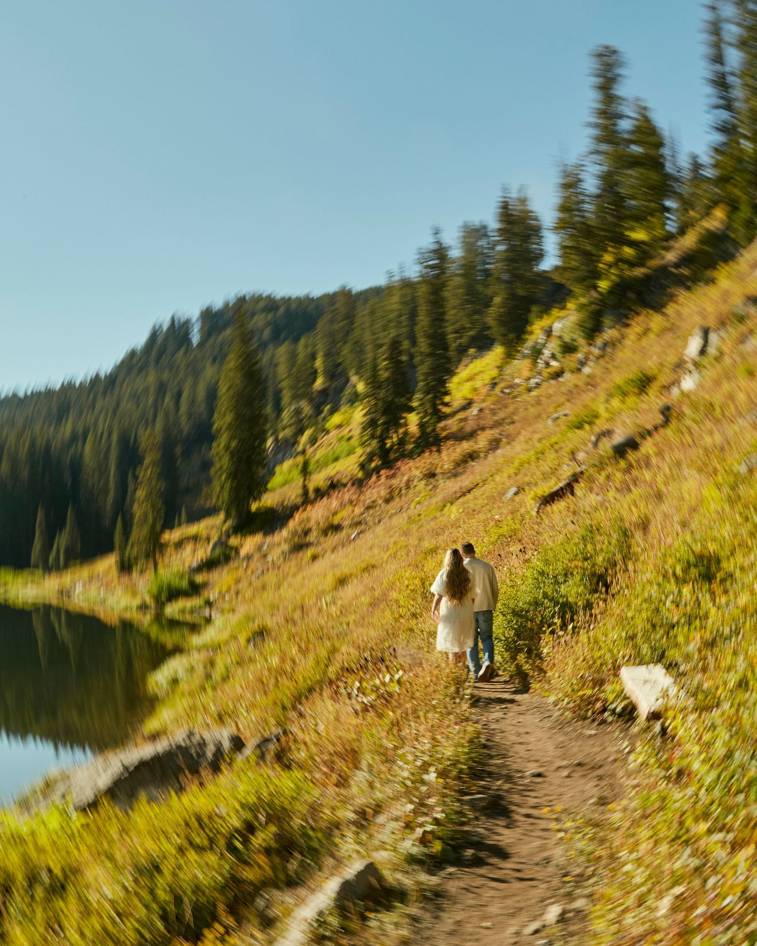 Person walking on mountain trail beside alpine lake with evergreen forest.