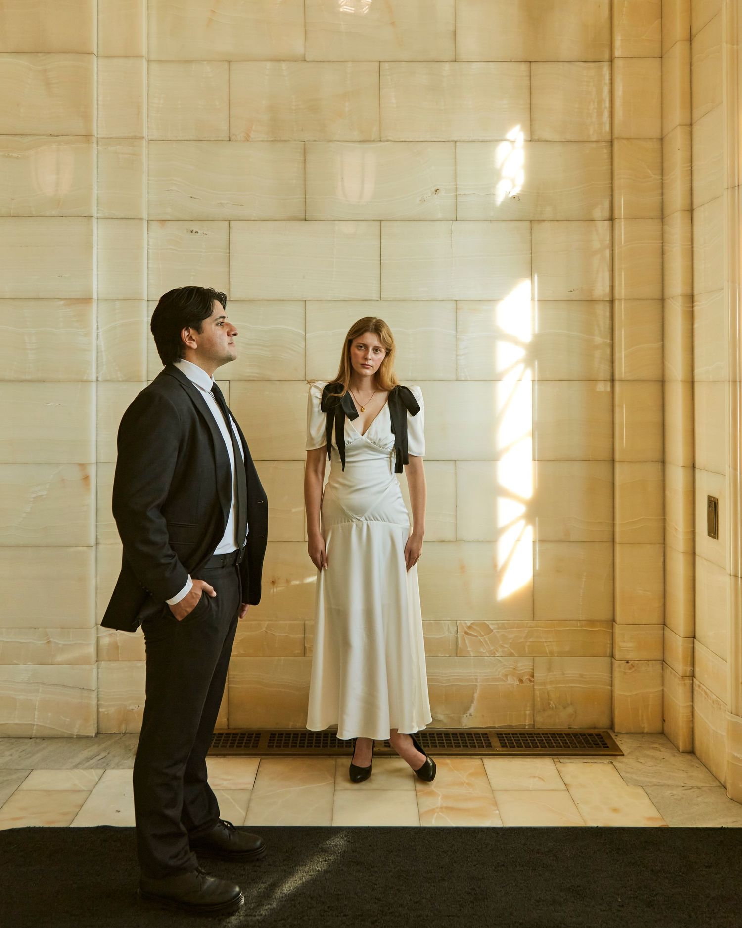 A couple in formal wear stands in a sunlit stone hallway.