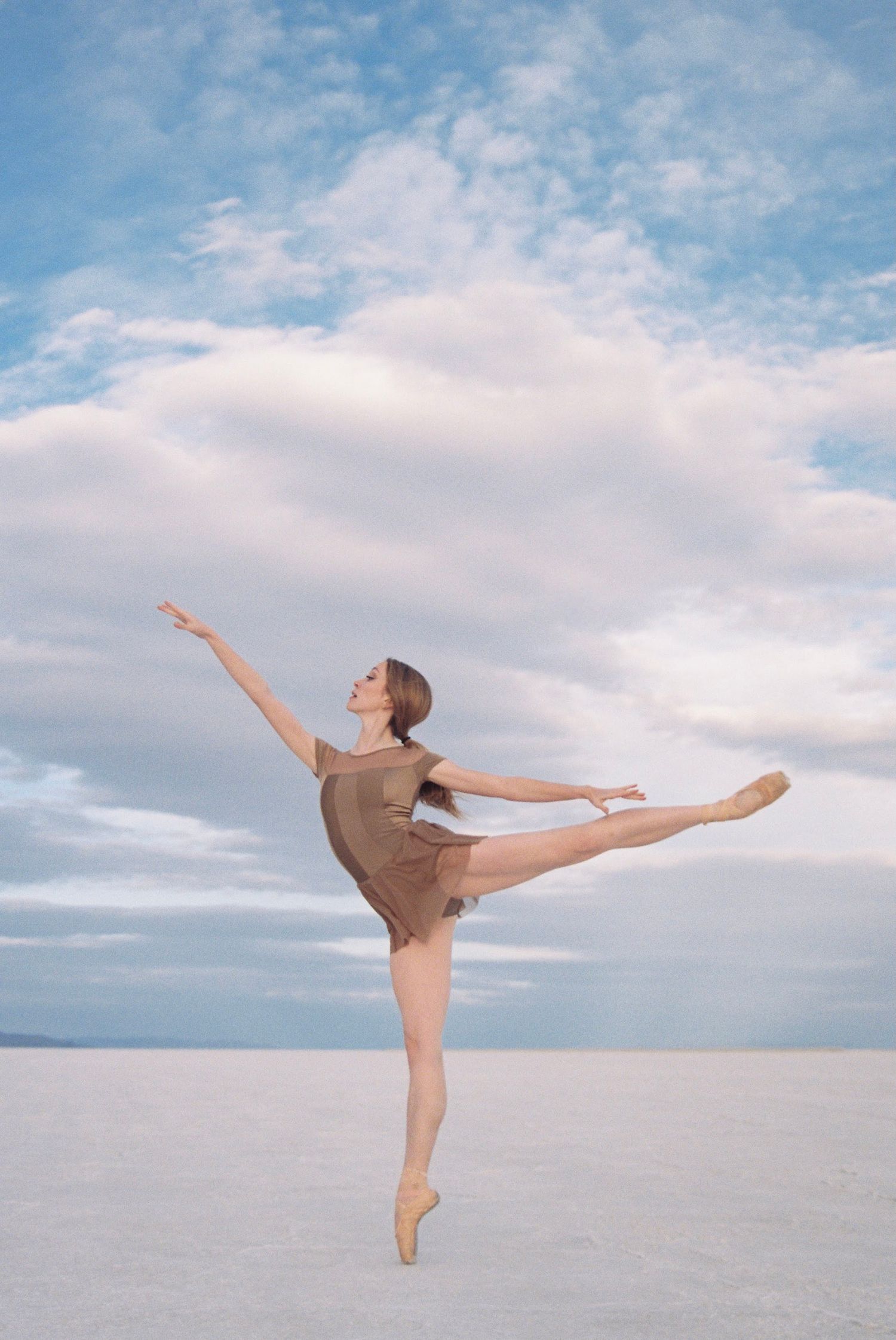 Ballet dancer performs an arabesque pose on white salt flats under cloudy skies.