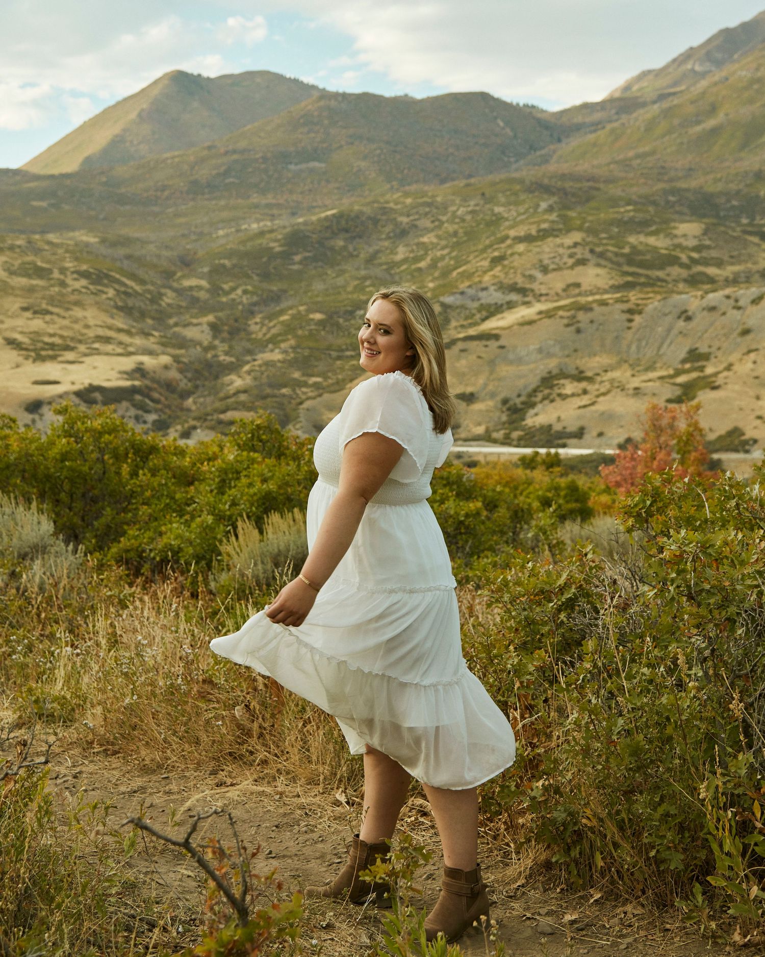 Person in flowing white dress poses against scenic mountain landscape on hiking trail.