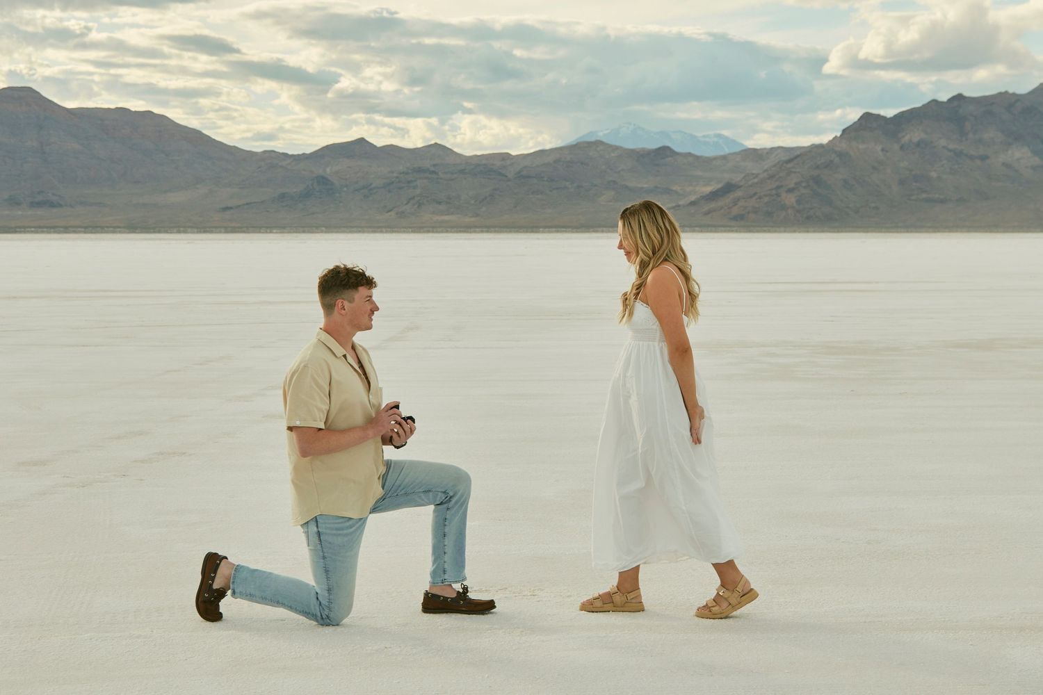 A romantic proposal scene on salt flats with mountain ranges and cloudy skies in the distance.