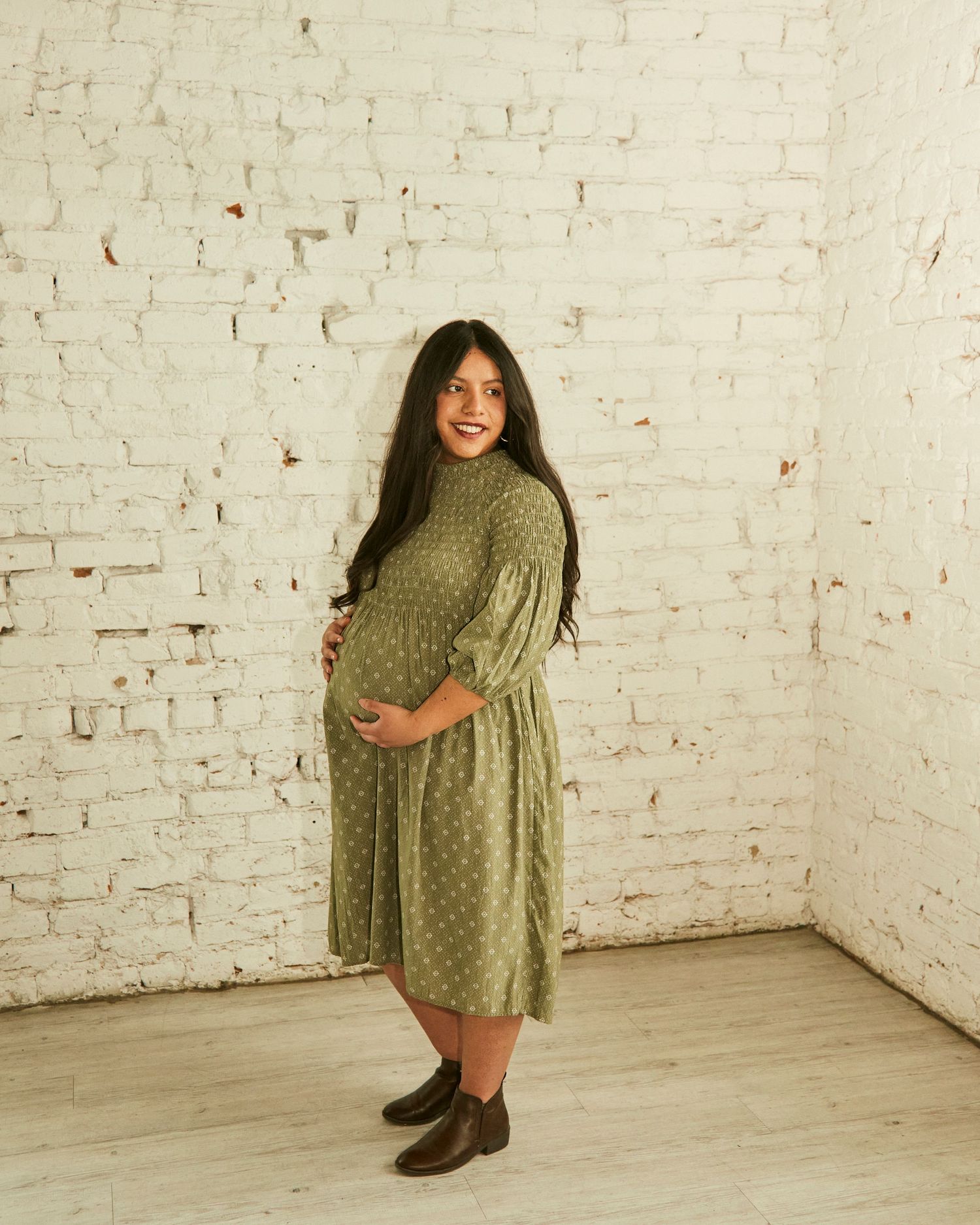 A green polka dot maternity dress is showcased against a white brick wall in a stylish indoor photoshoot.