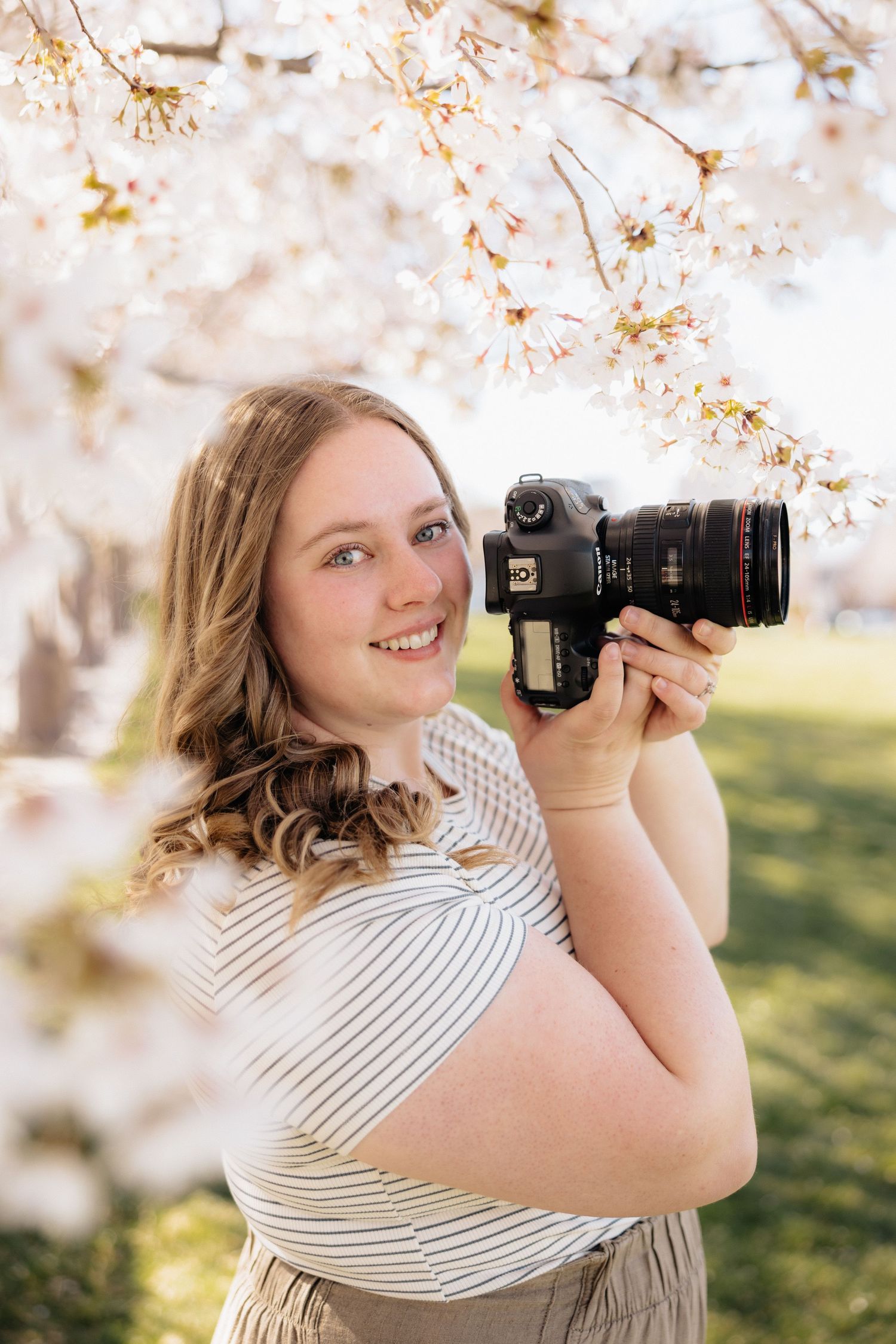 A photographer with a Canon camera captures spring blossoms under flowering cherry trees in soft natural light.