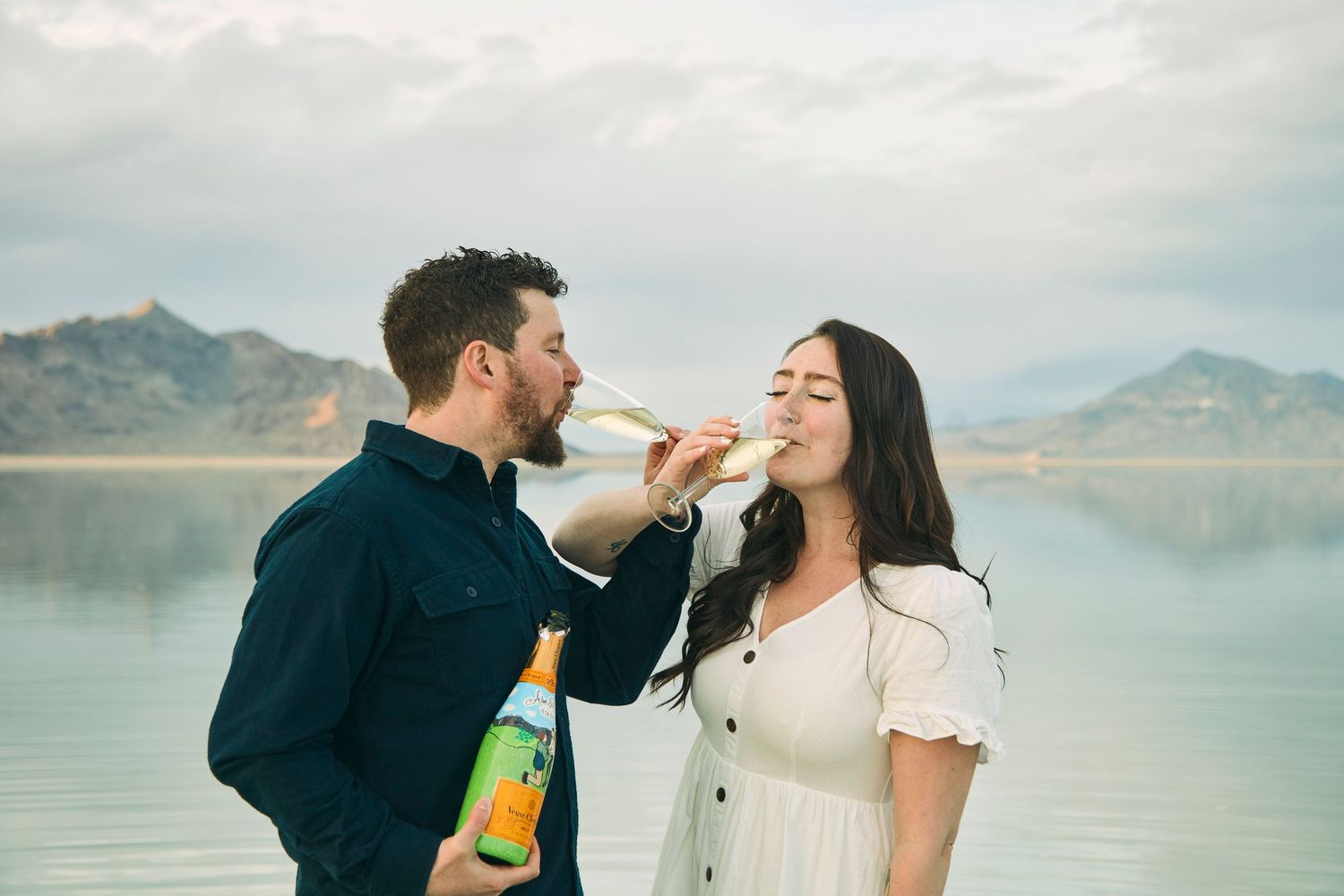 Playful couple sharing champagne by a scenic mountain lake.