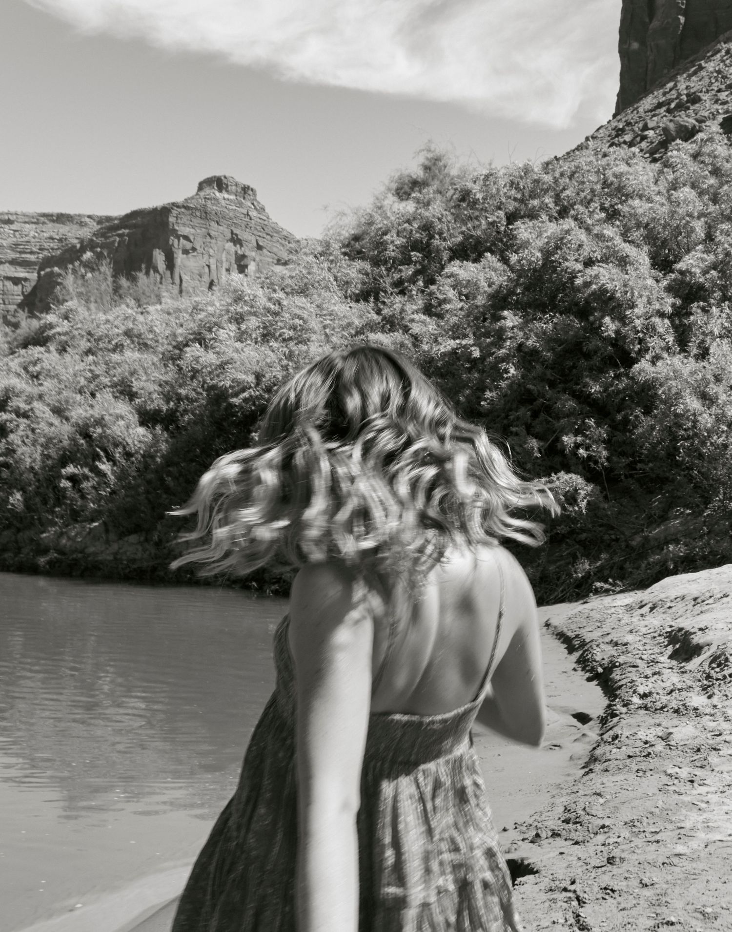 Black and white photo of a dress-clad figure with flowing hair by a lake and mountains.