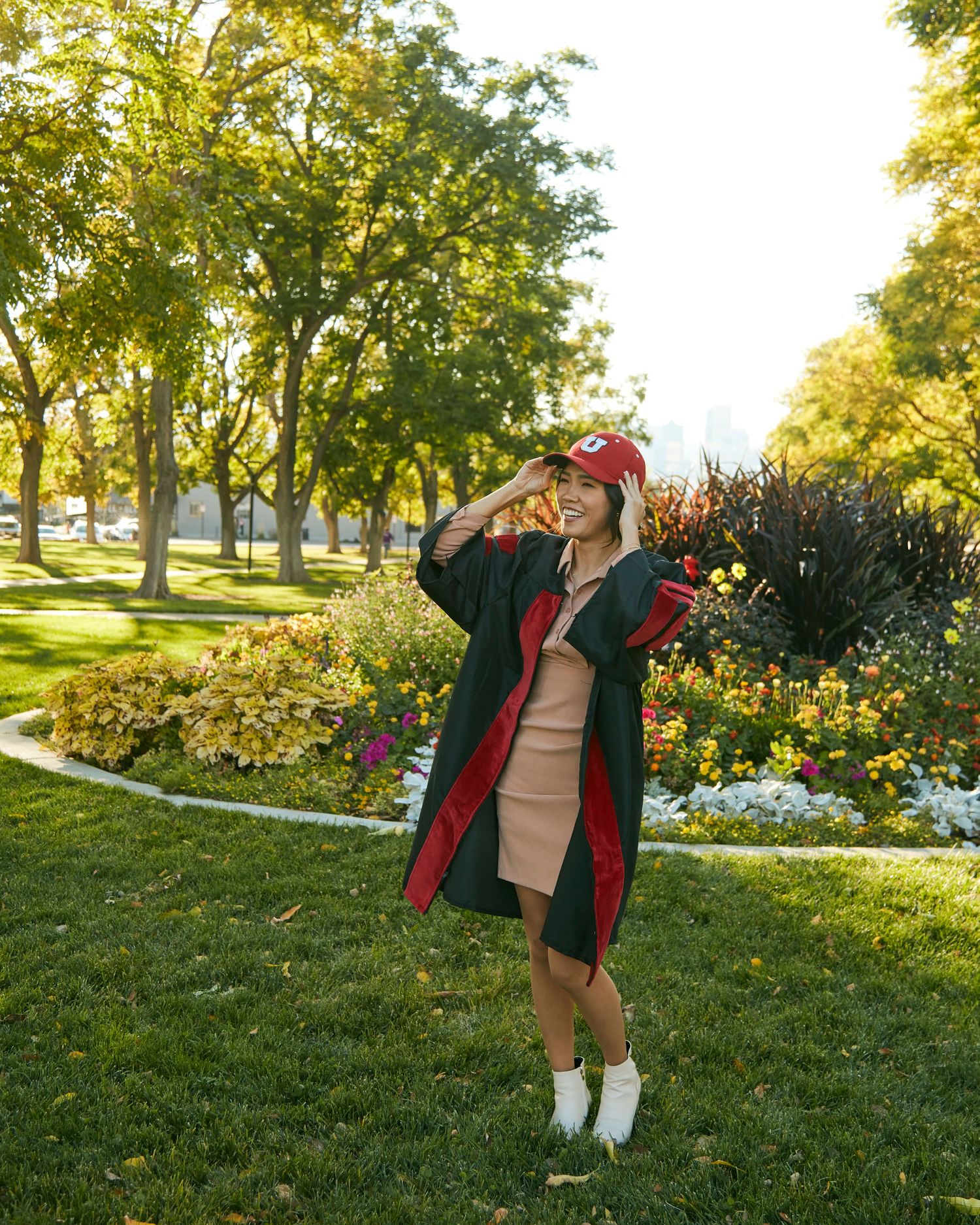 A graduate in cap and gown poses joyfully in a sunny park with trees and flowers.