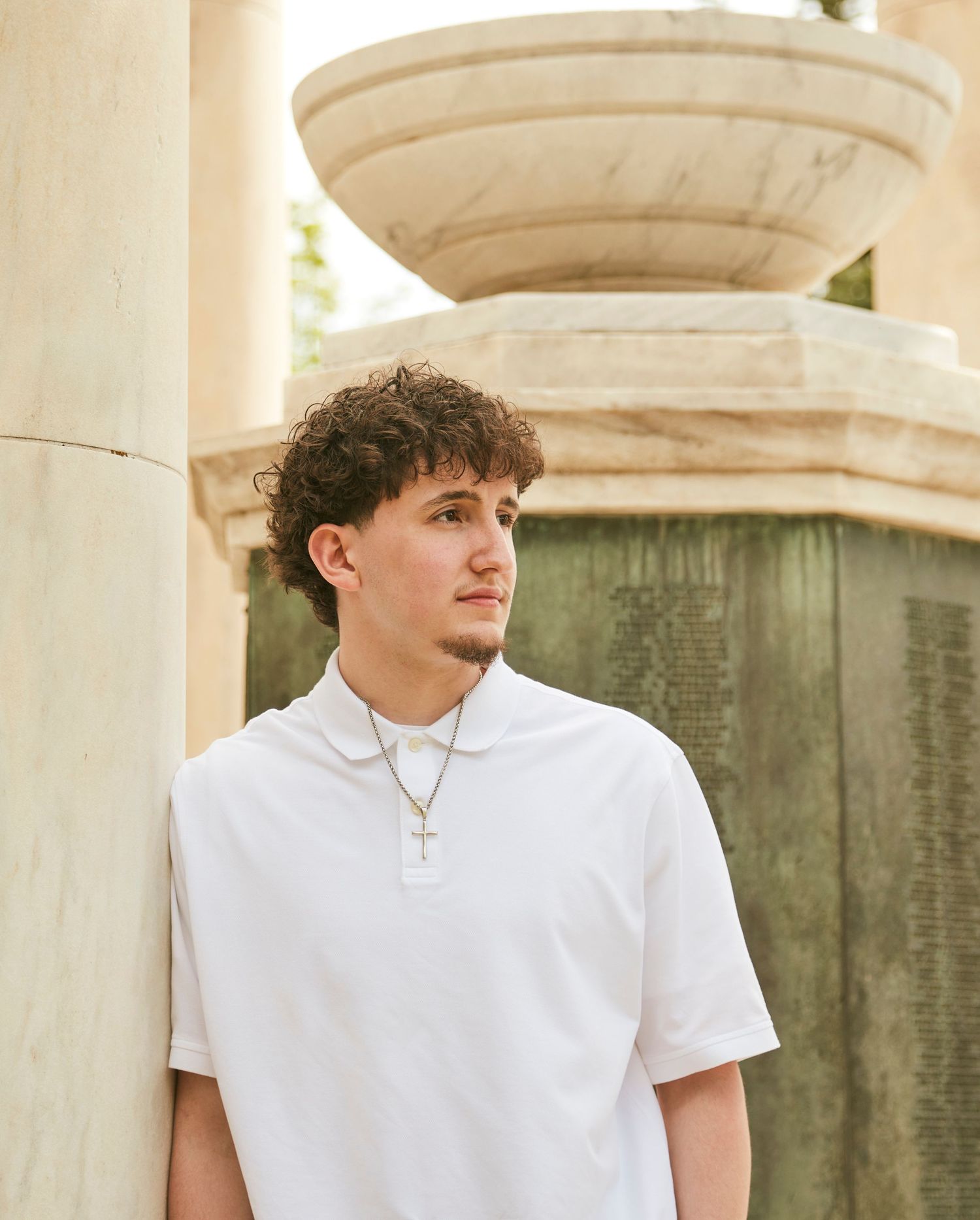 Portrait against stone columns showing person in white polo shirt with necklace.
