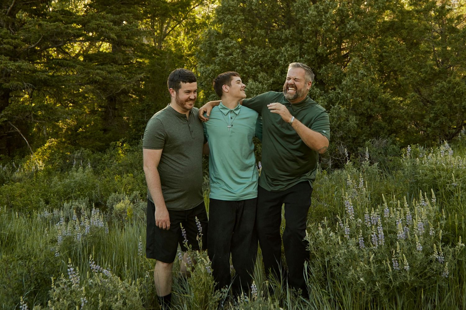 Three friends sharing laughter in a meadow with evergreen trees at sunset.