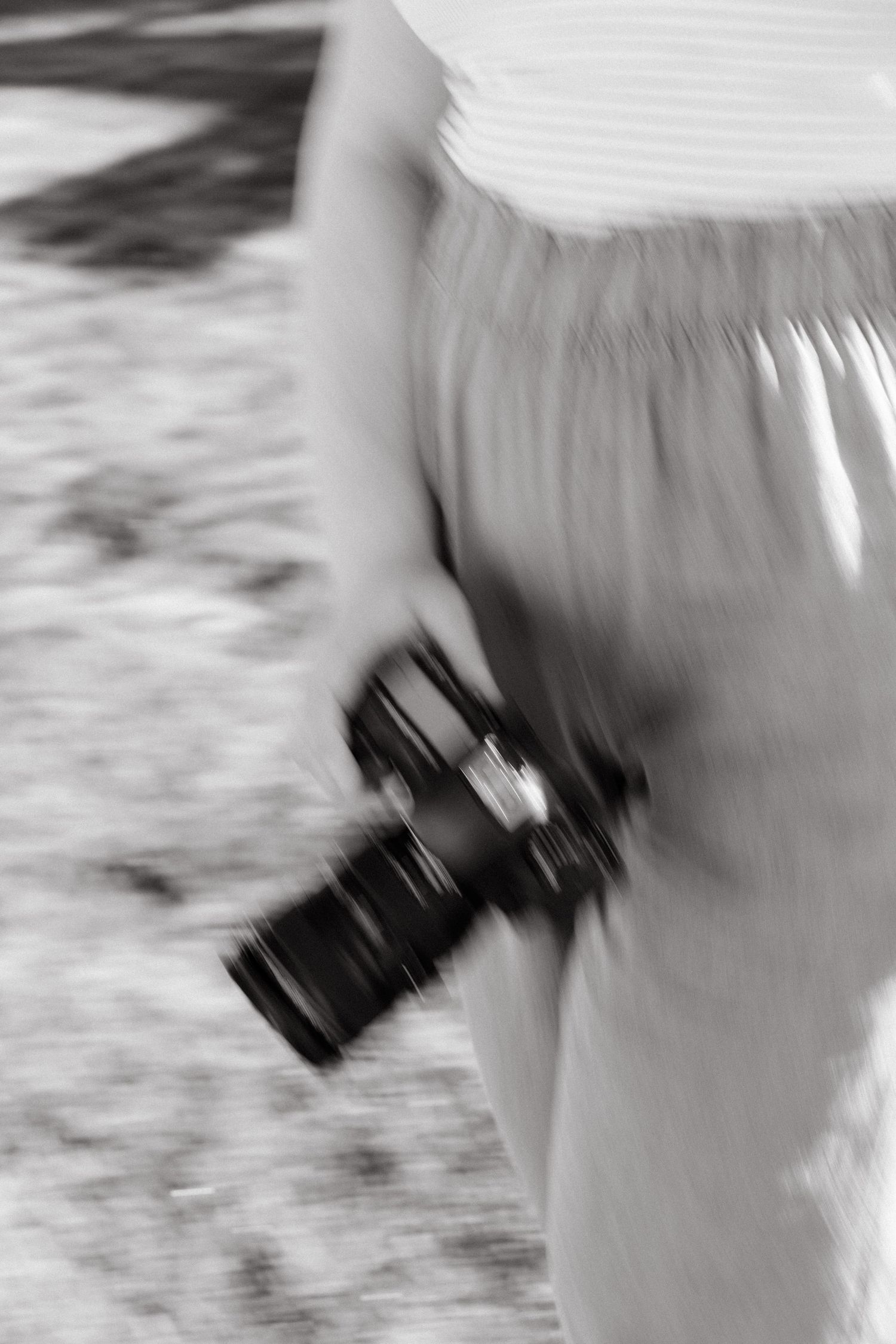 A camera hangs at someone's side against a white flowing dress, captured in artistic black and white motion blur.