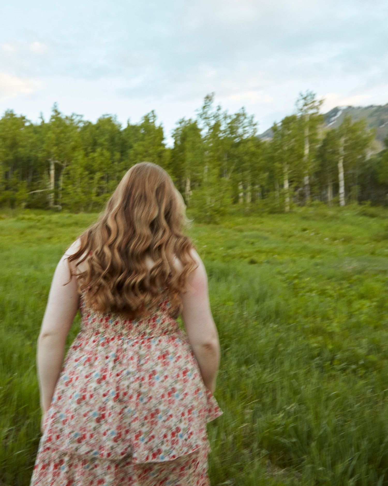 Person with wavy hair in floral dress looking out at green mountain meadow.
