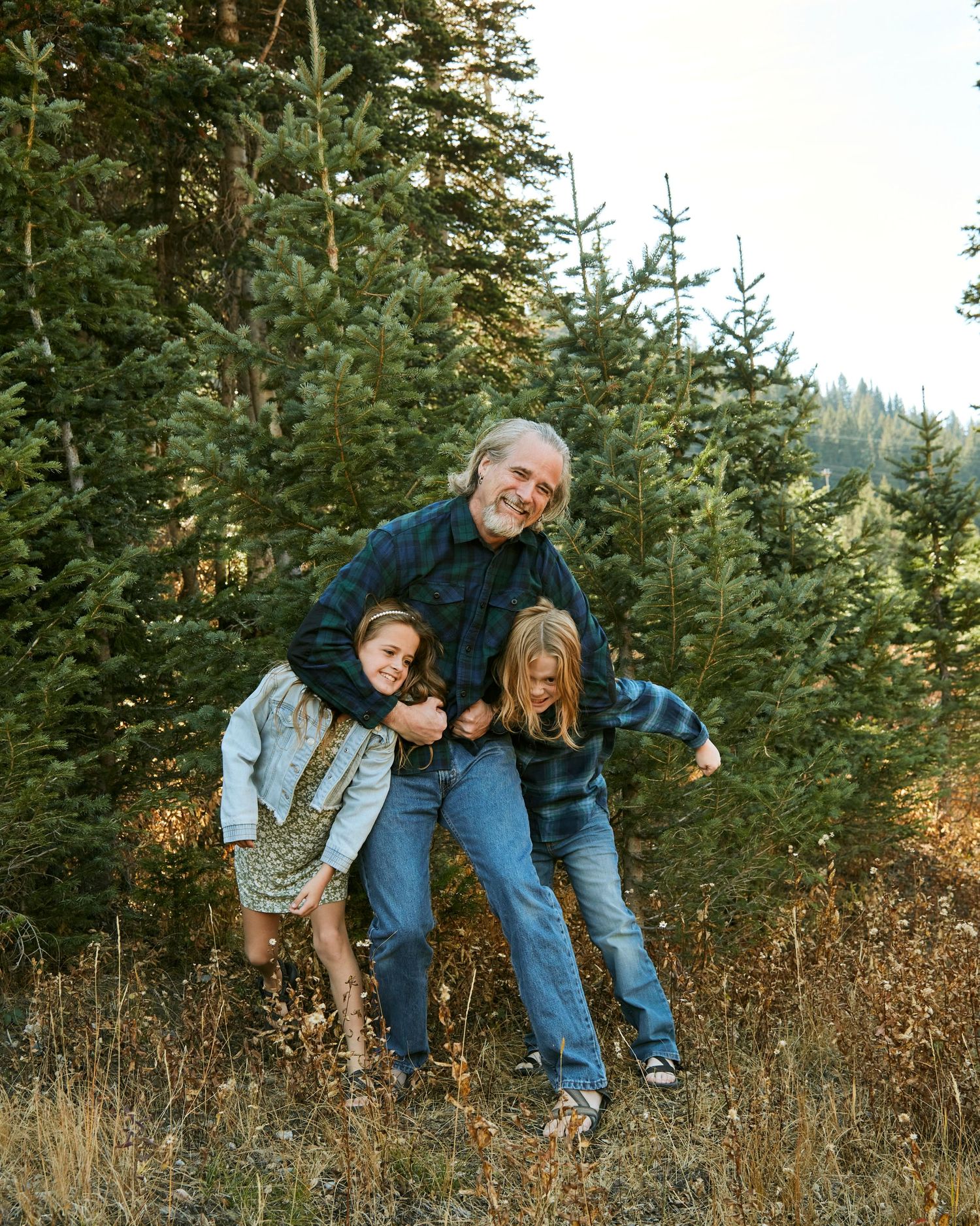 Playful outdoor family moment captured between evergreen trees in casual denim outfits.