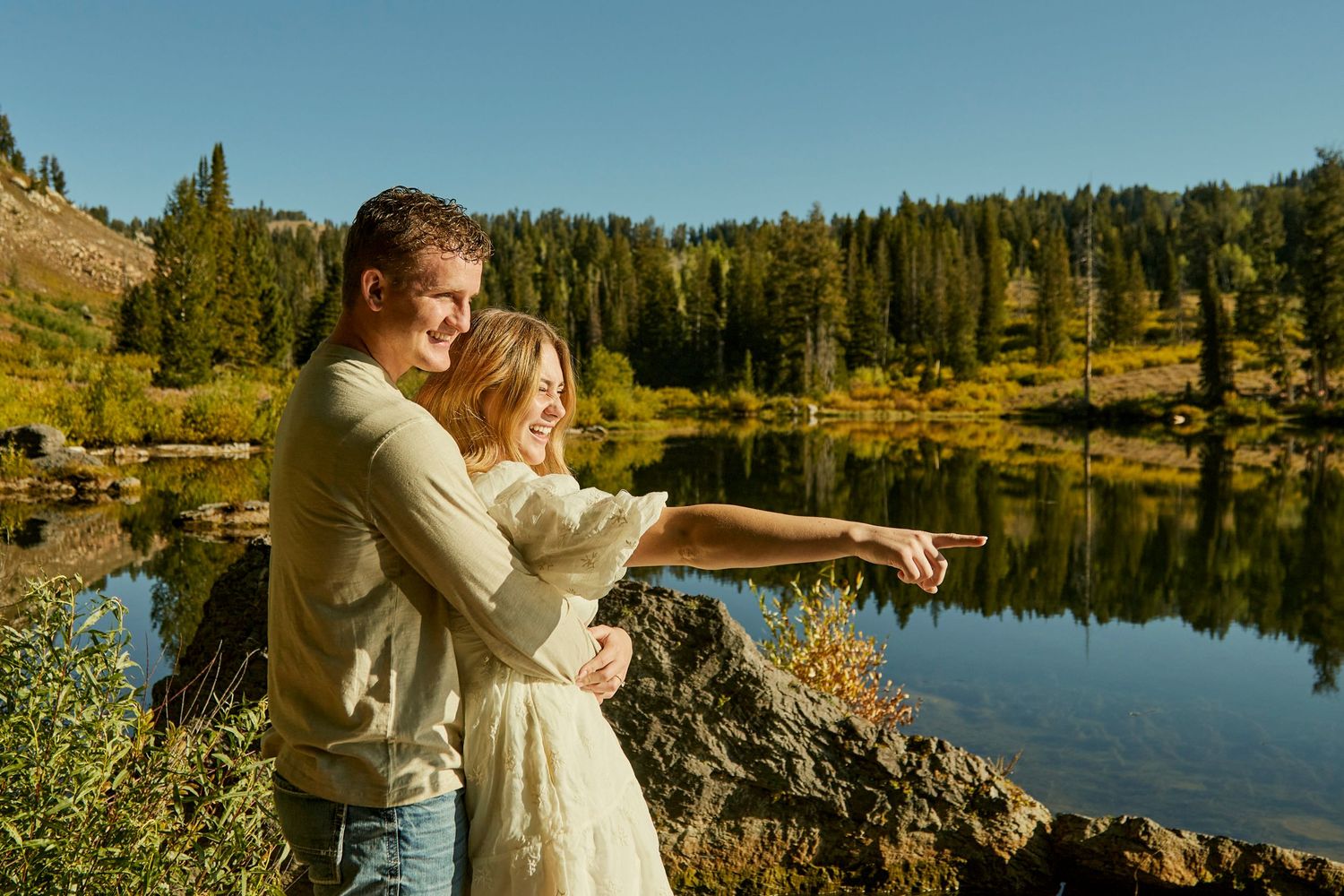 Two people sharing a romantic moment by a serene mountain lake with autumn foliage and forest scenery.