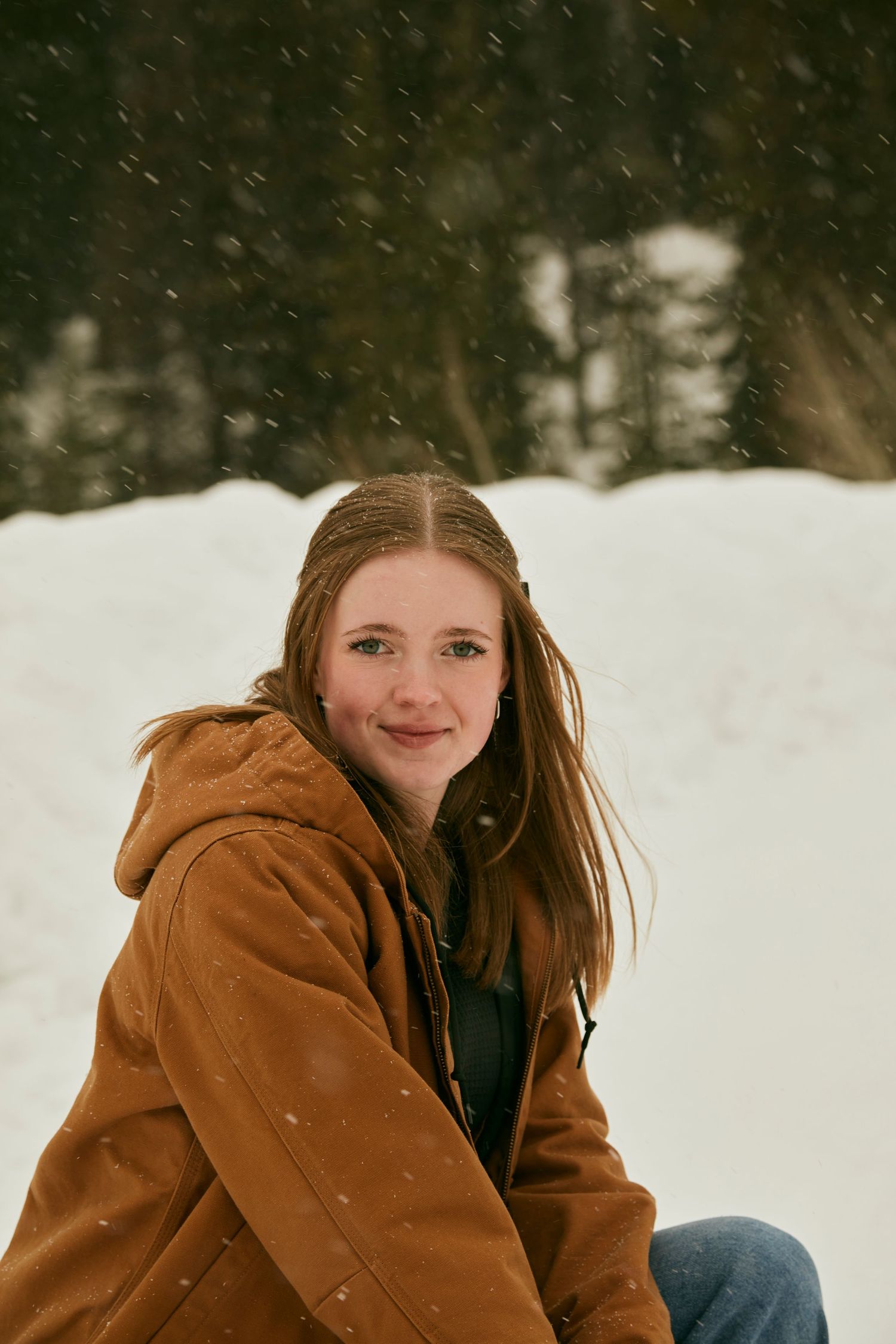 Person in brown jacket sits in snowy winter landscape with falling snowflakes.
