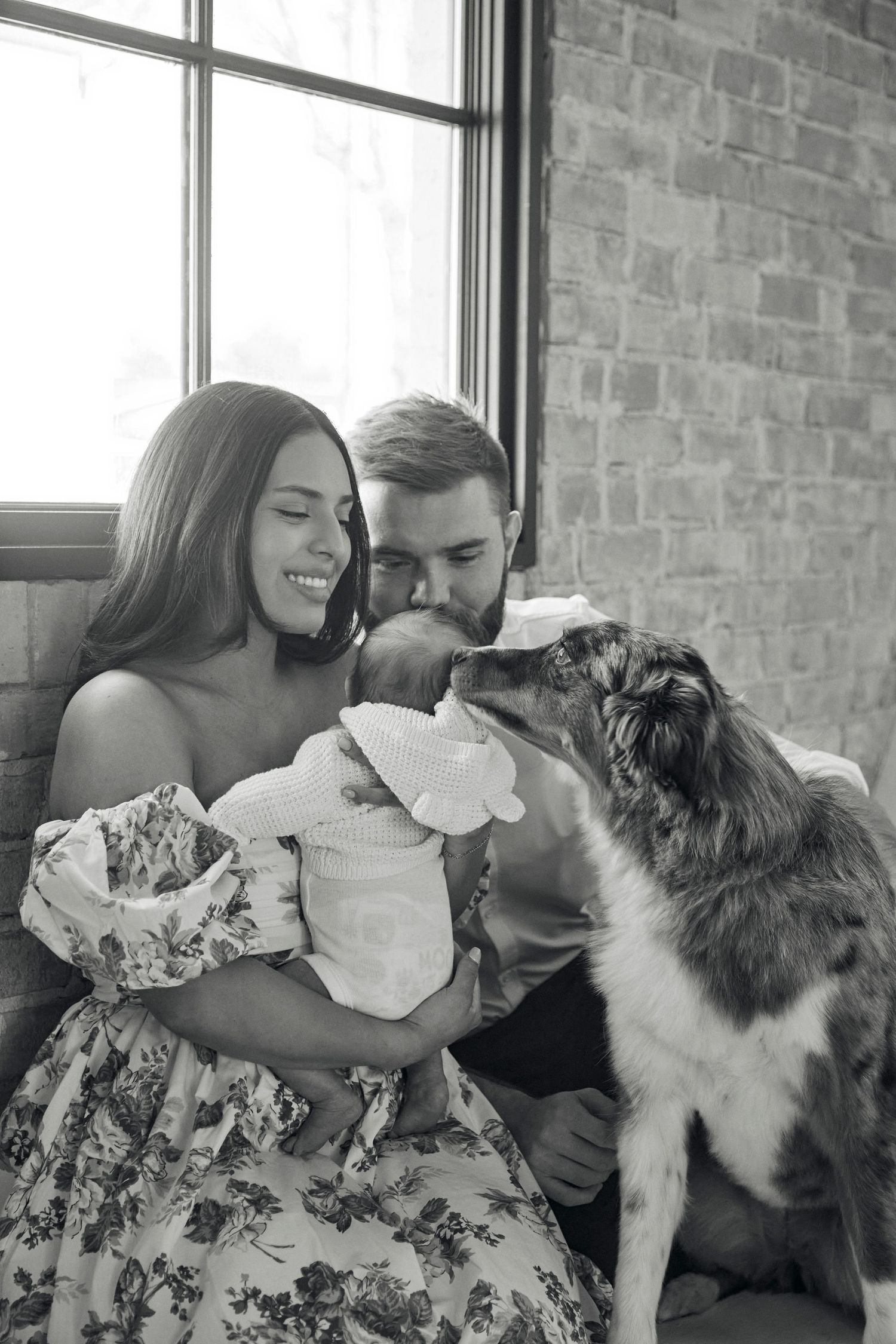 Family and dog share tender moment with newborn baby by bright window.