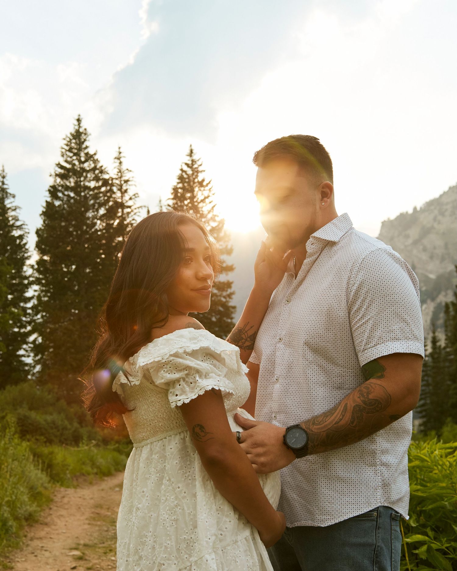Couple in white clothing embracing during golden hour with mountain scenery and evergreen trees in background.