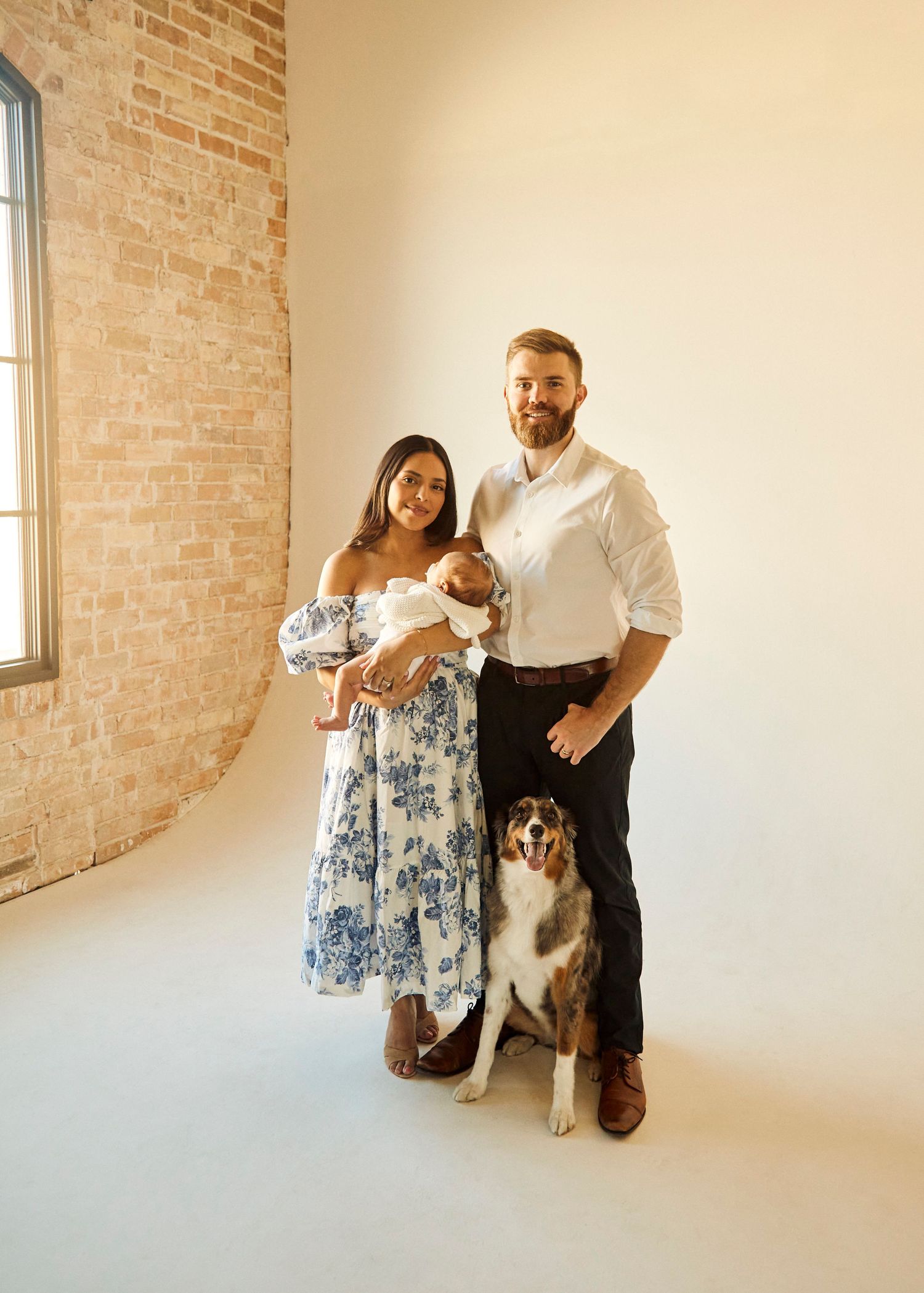 Family portrait in a sunny room with exposed brick wall featuring a baby, couple, and Australian shepherd dog.