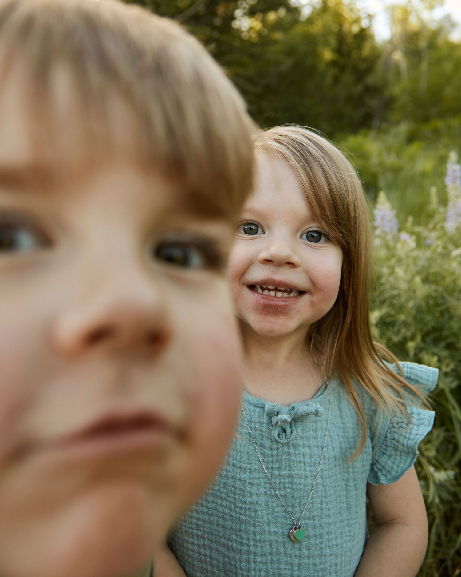 Close-up playful portrait of children in a field with one face in focus and another blurred.