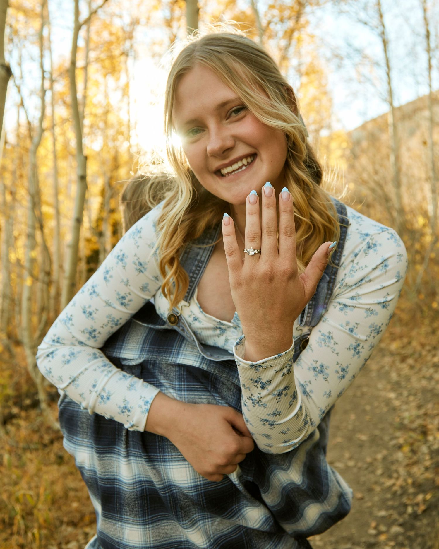Person in overalls and floral top showing off engagement ring in autumn forest setting.