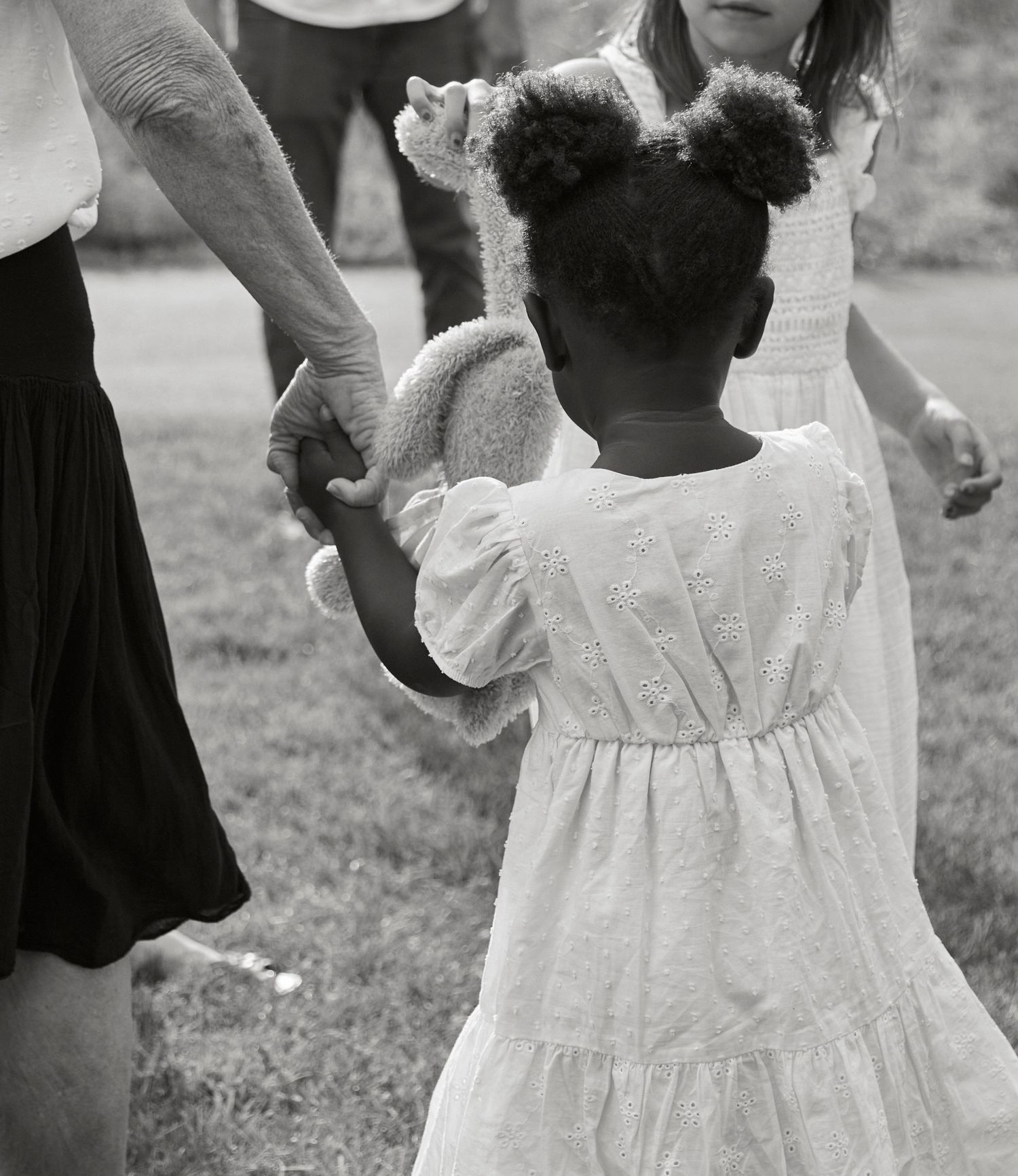 Black and white image of a child in white dress holding hands with adults outdoors.