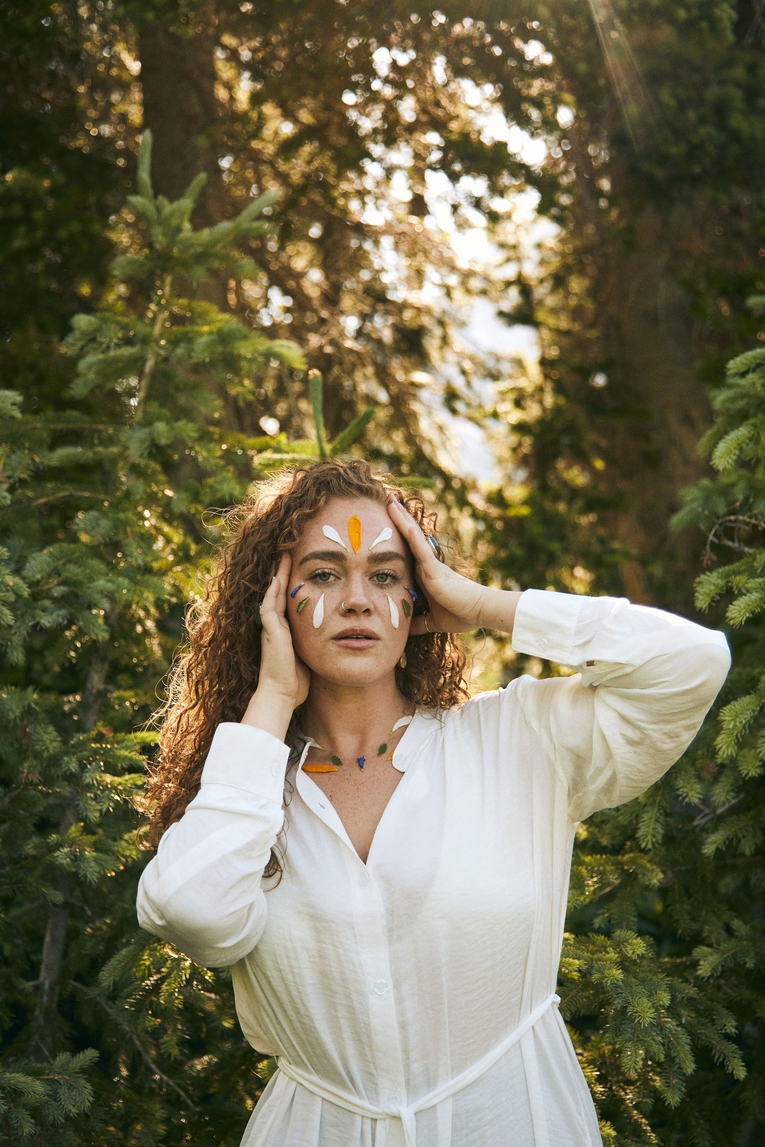 Ethereal portrait with decorative face paint surrounded by green forest foliage.