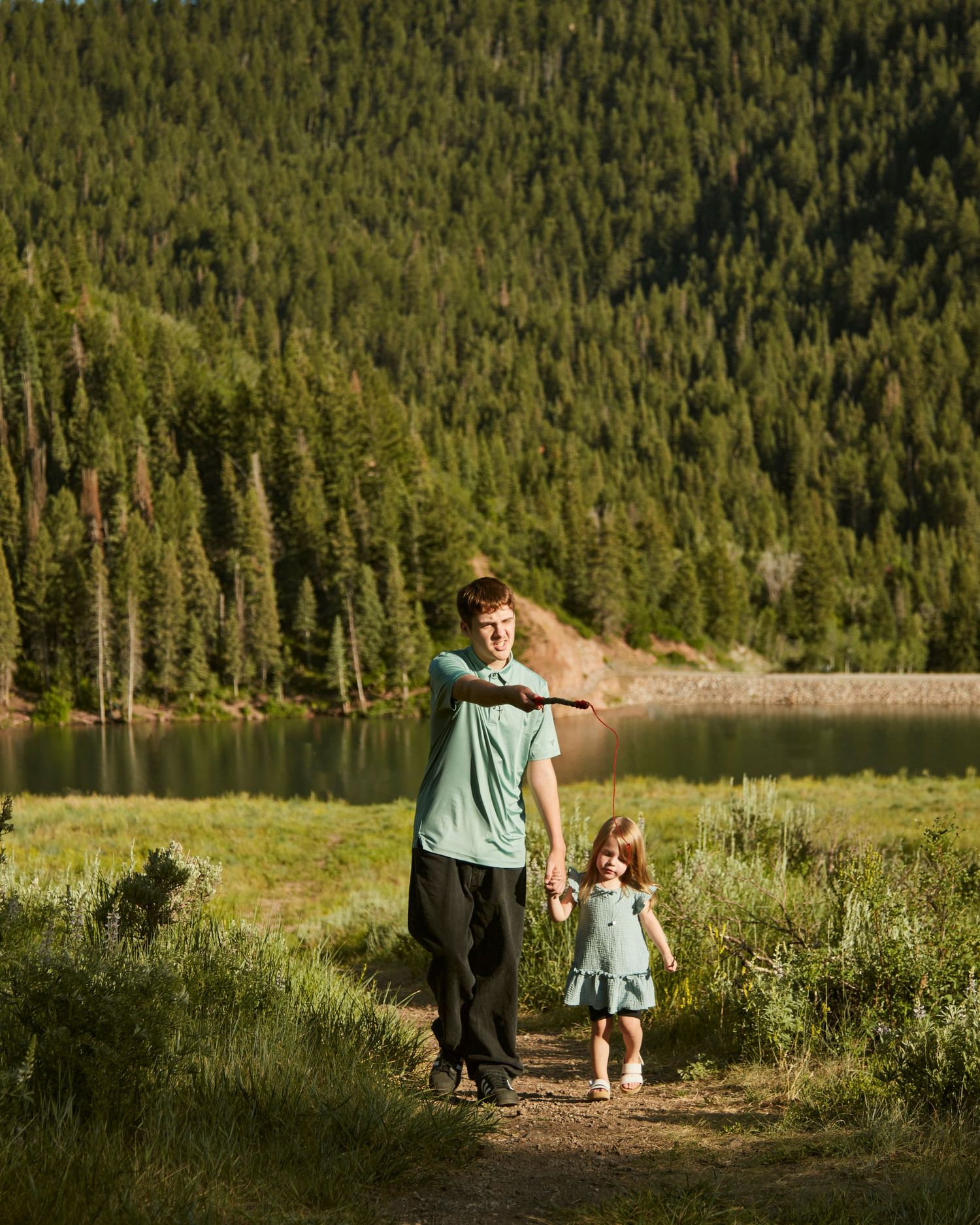 A pair walking along a serene mountain lake surrounded by dense pine forest.