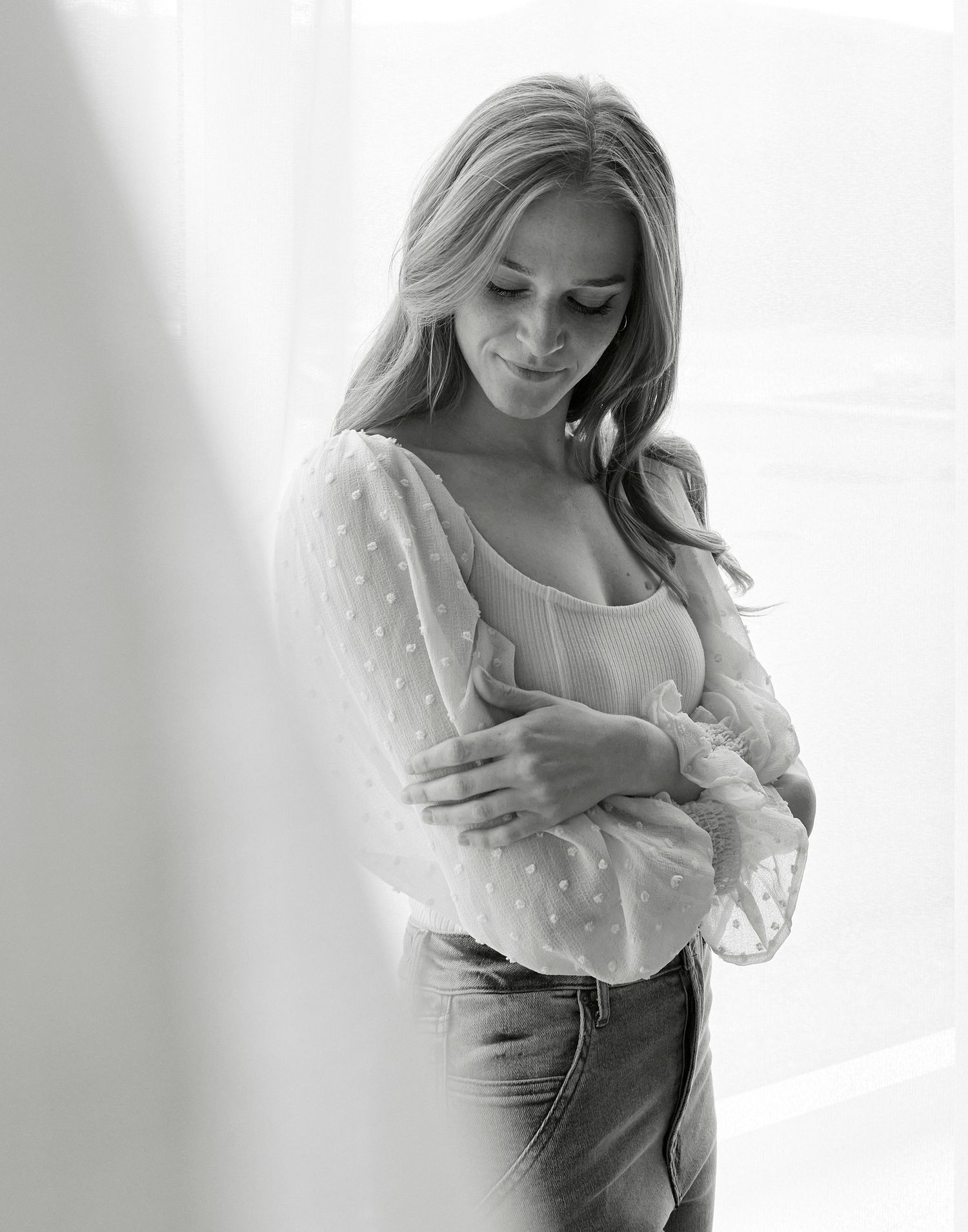 A black and white portrait of a person in a sheer blouse with delicate details posing against a light background.