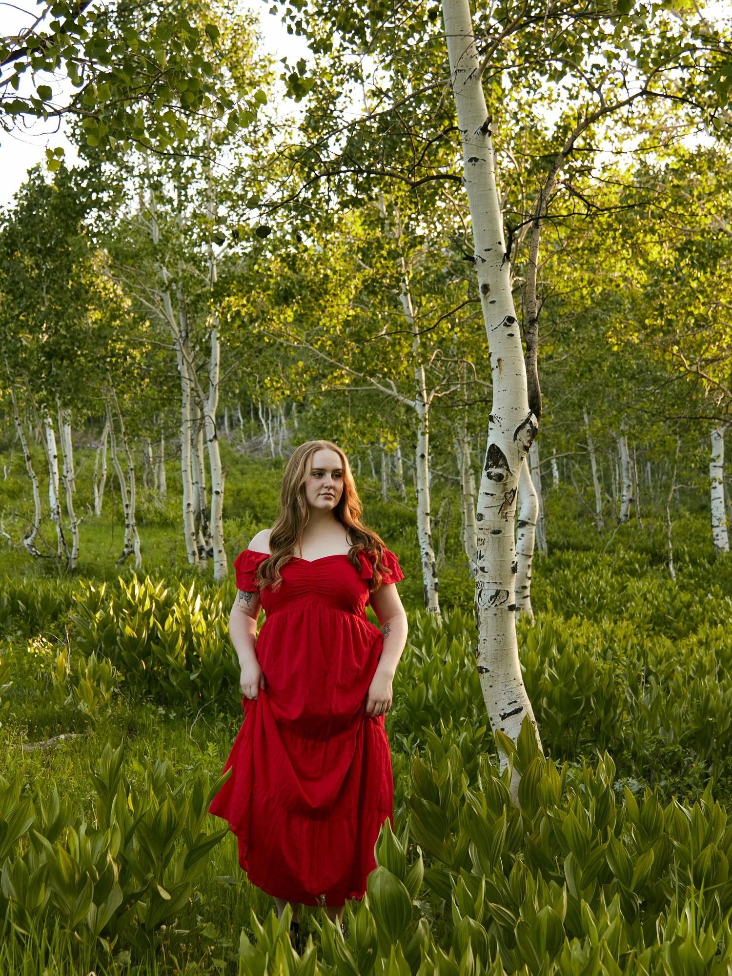 A person in a bright red dress standing among tall birch trees in a lush green meadow.
