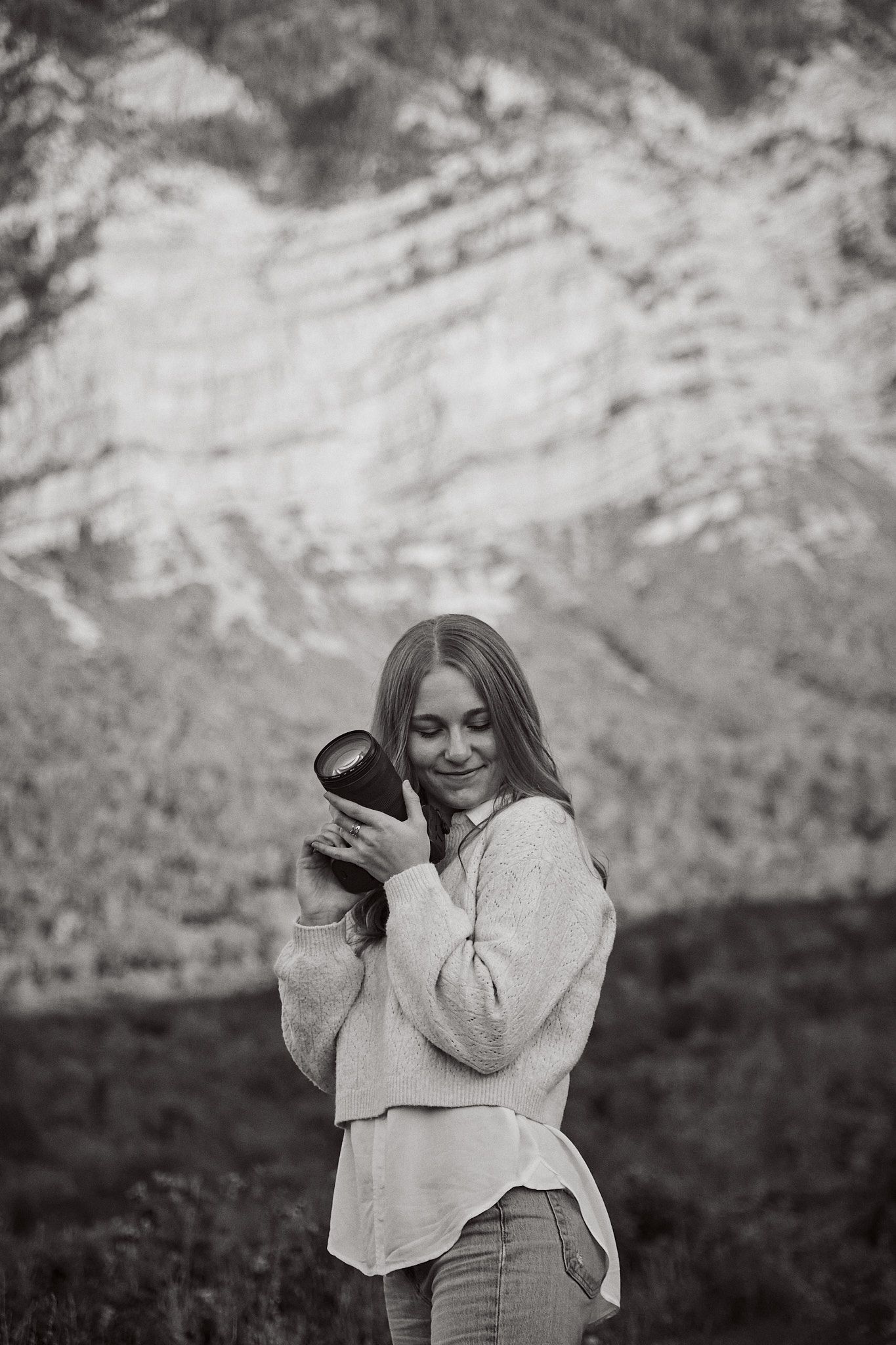Black and white photo with mountain backdrop showing person holding camera in cozy sweater.