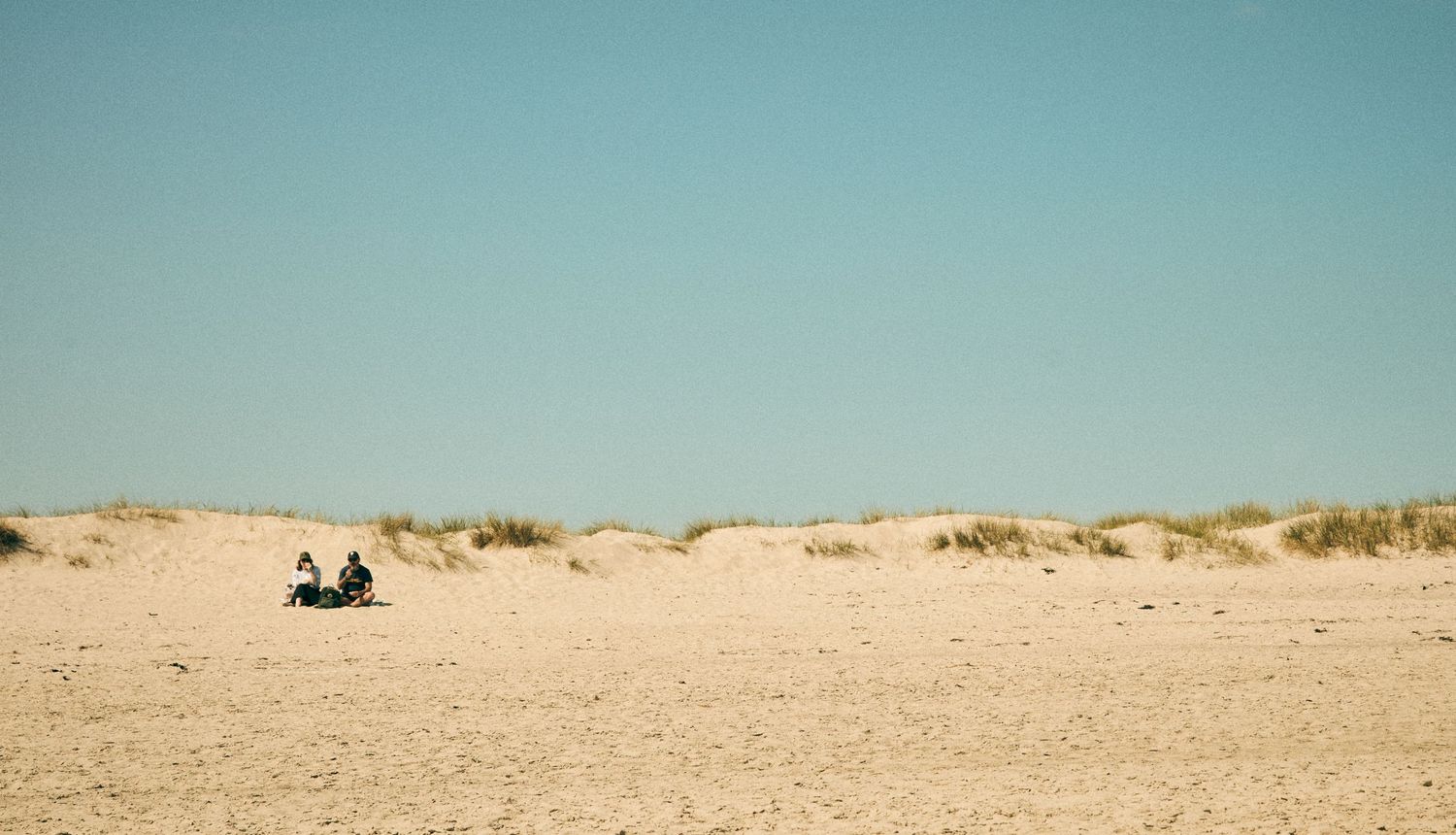 Two distant figures sitting on vast sandy beach with dunes in background.