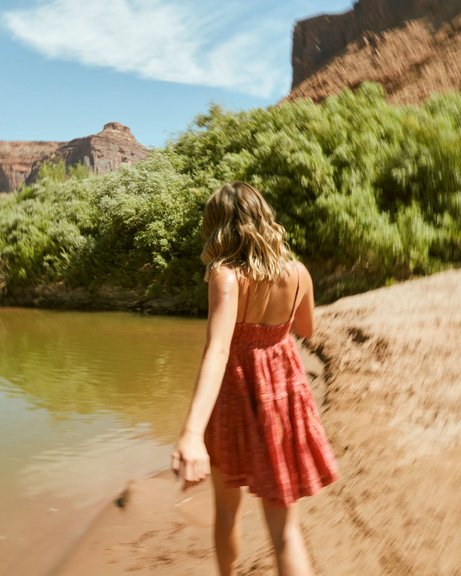 A summery coral dress catches sunlight on a sandy riverbank with towering canyon walls.