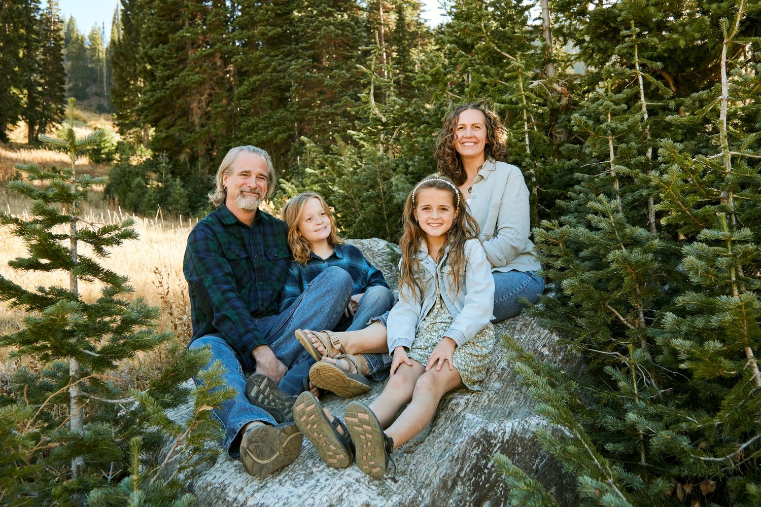 A family sitting together on a rock surrounded by evergreen trees in a mountain setting.