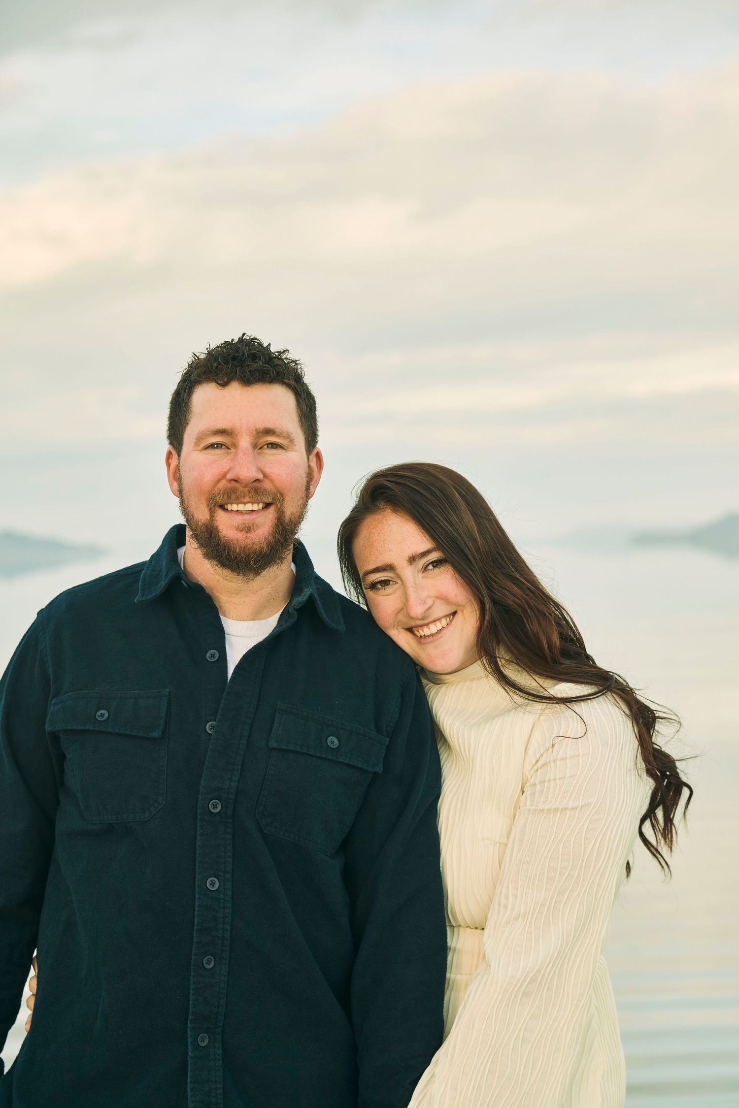 Couple in casual winter wear smiling against scenic mountain lake backdrop.