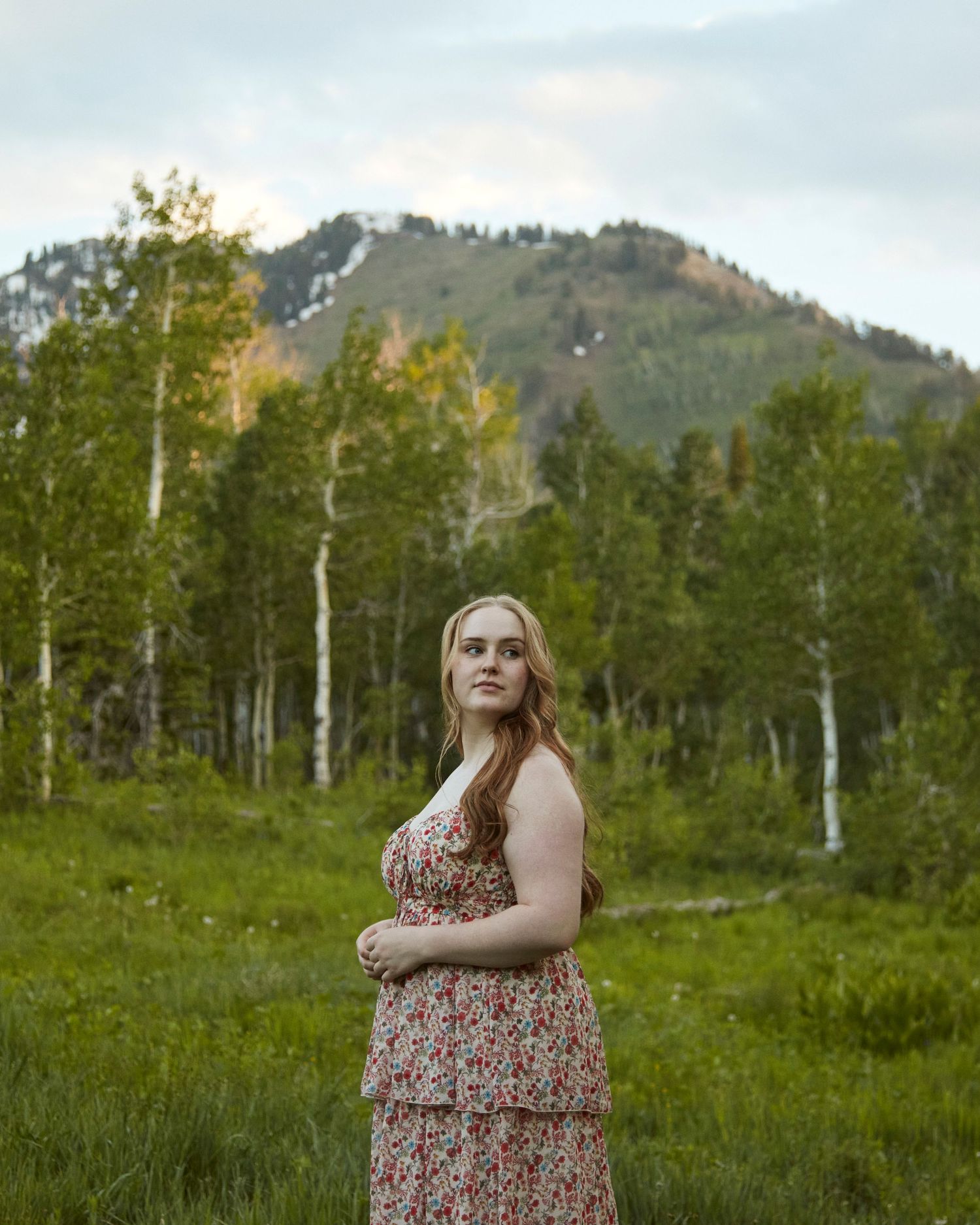 A person in a floral dress poses in a mountain meadow with aspen trees and hills in the background.