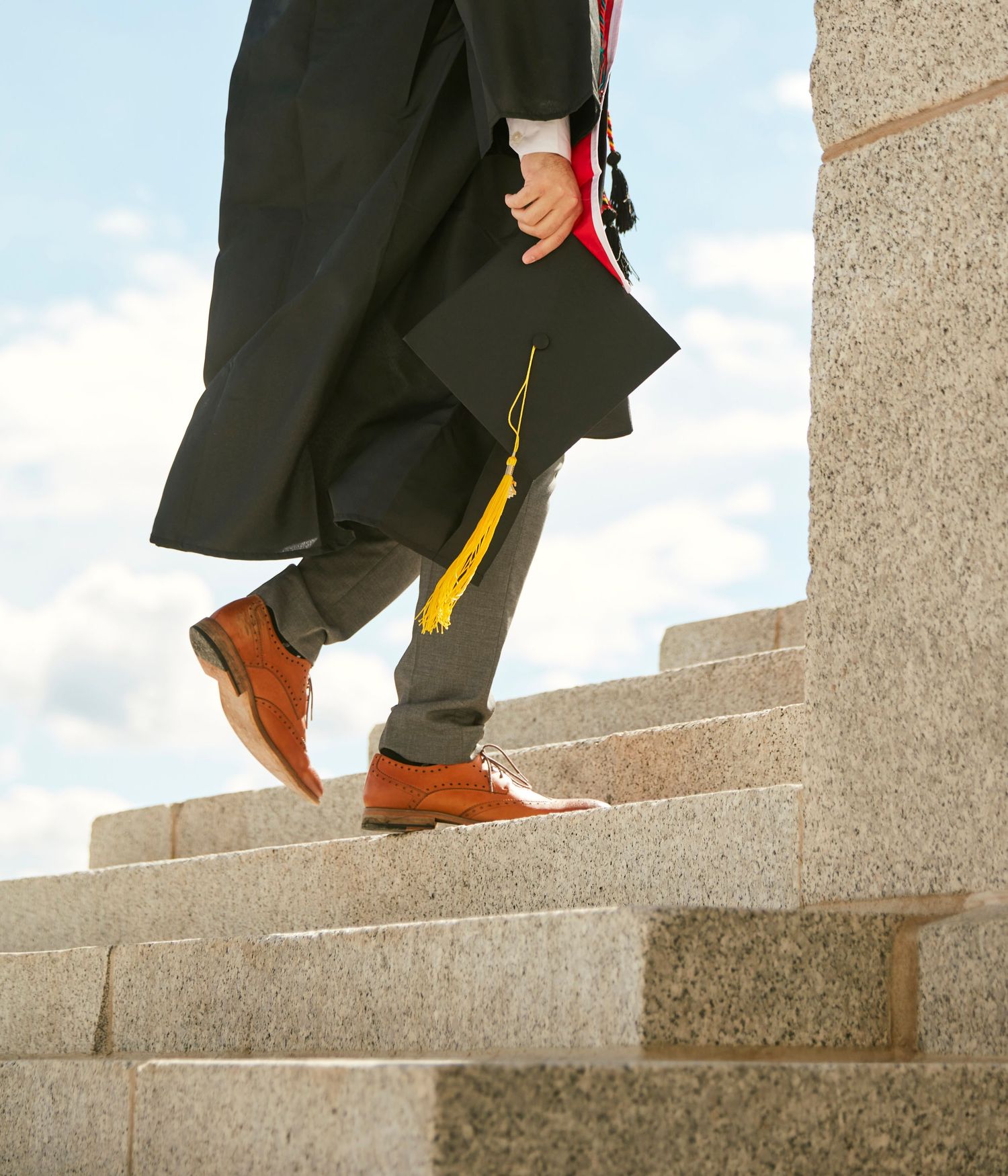 Close-up of graduation robe and brown shoes walking up concrete steps against blue sky.