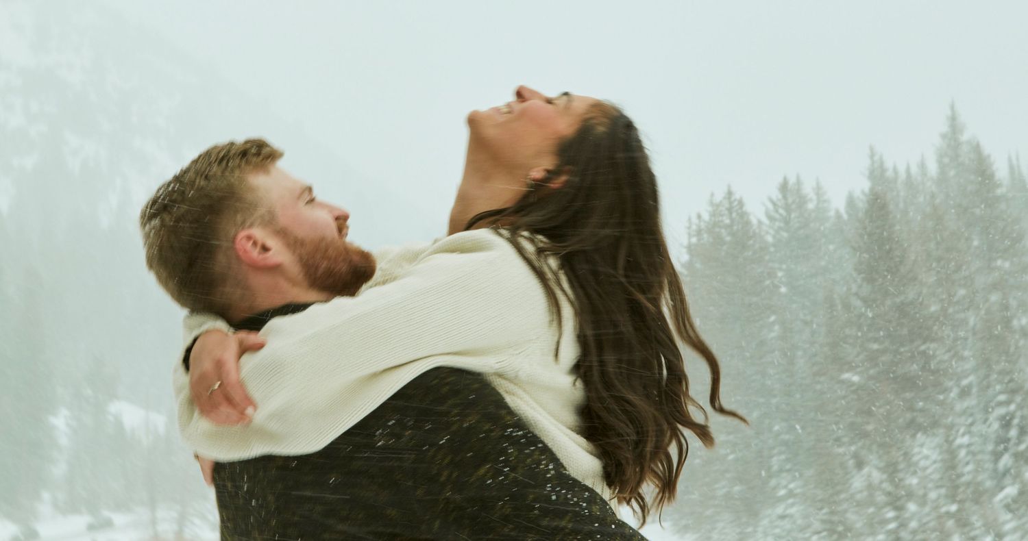 Couple sharing playful moment in winter wonderland with snow-covered pine trees in background.