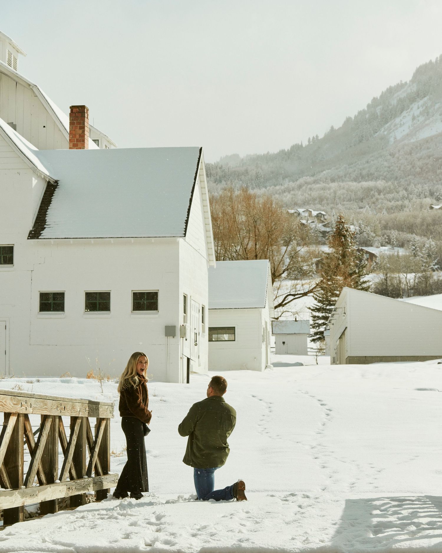 Two people stand in deep snow near white buildings with mountains rising in the background.