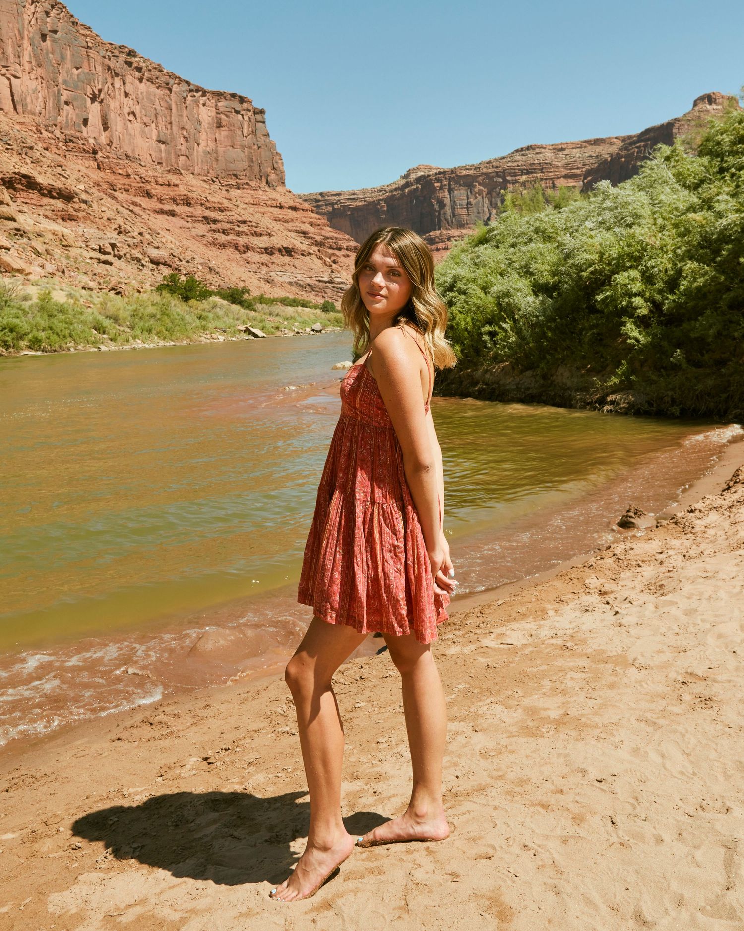 Carefree pose in coral dress along desert riverbank with red rock formations.