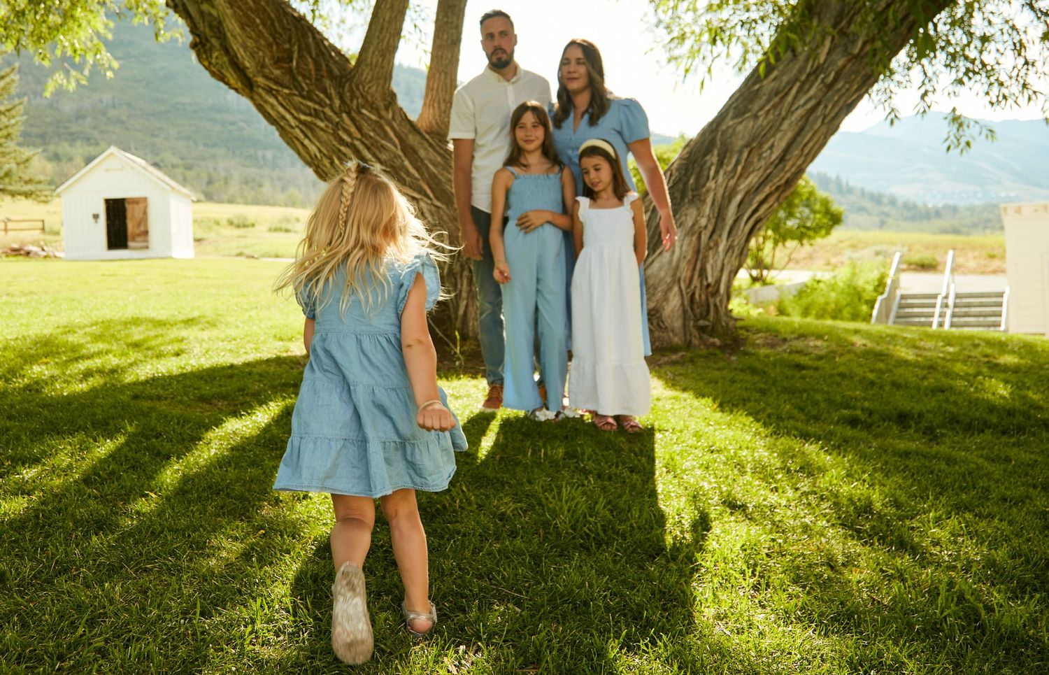 A casual outdoor family photoshoot under a large shaded tree with mountains and a white building in the background.
