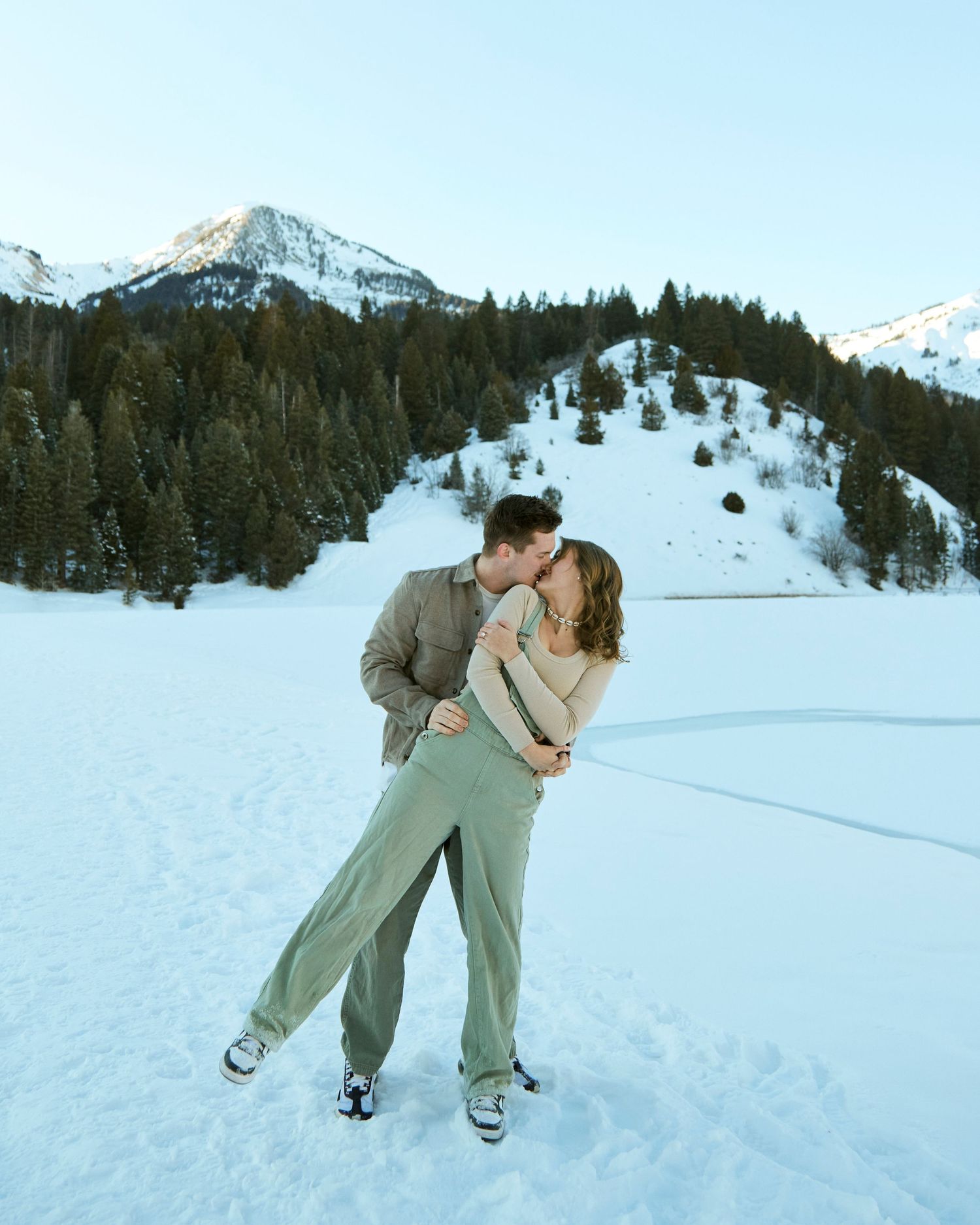 Couple embracing in front of snow-covered mountains and evergreen forest on a winter afternoon.