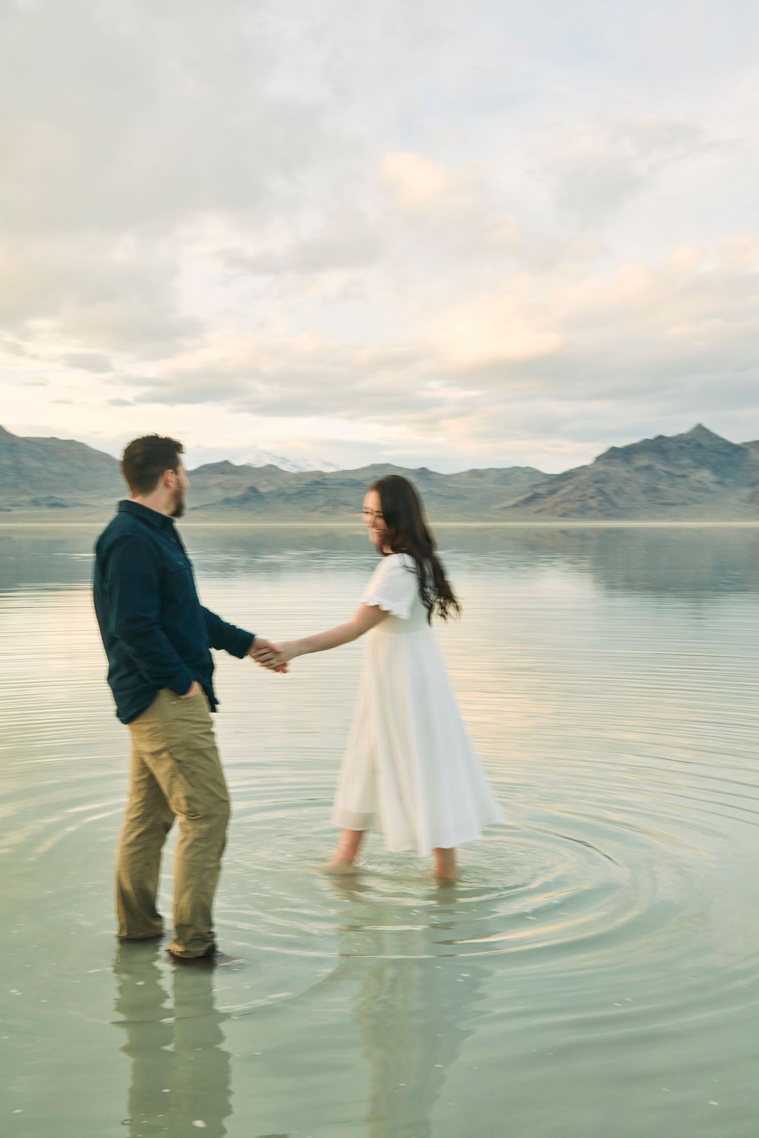 Couple holding hands while standing in shallow reflective water with mountains in background.