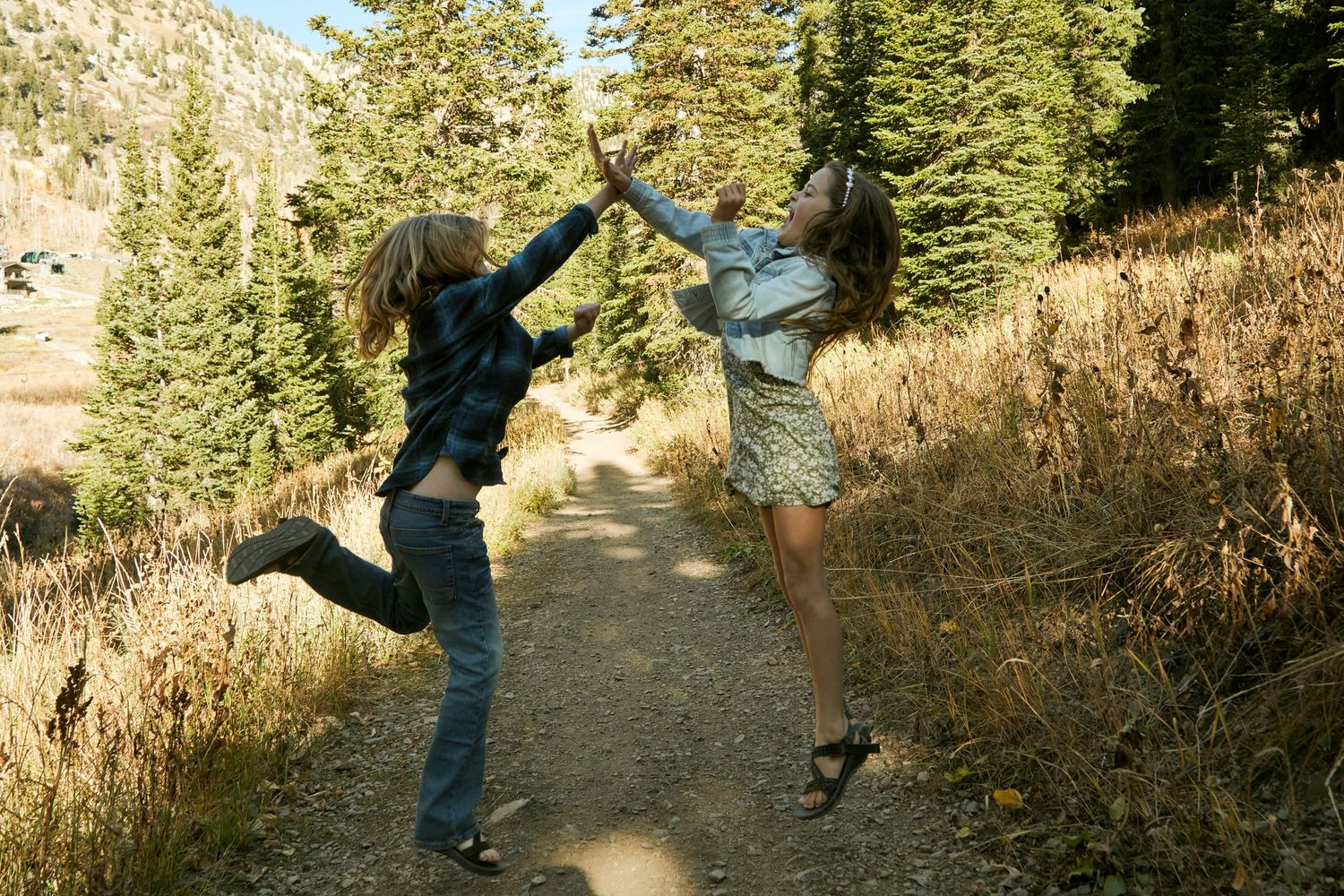 Two people jumping and high-fiving on a mountain trail surrounded by evergreens.
