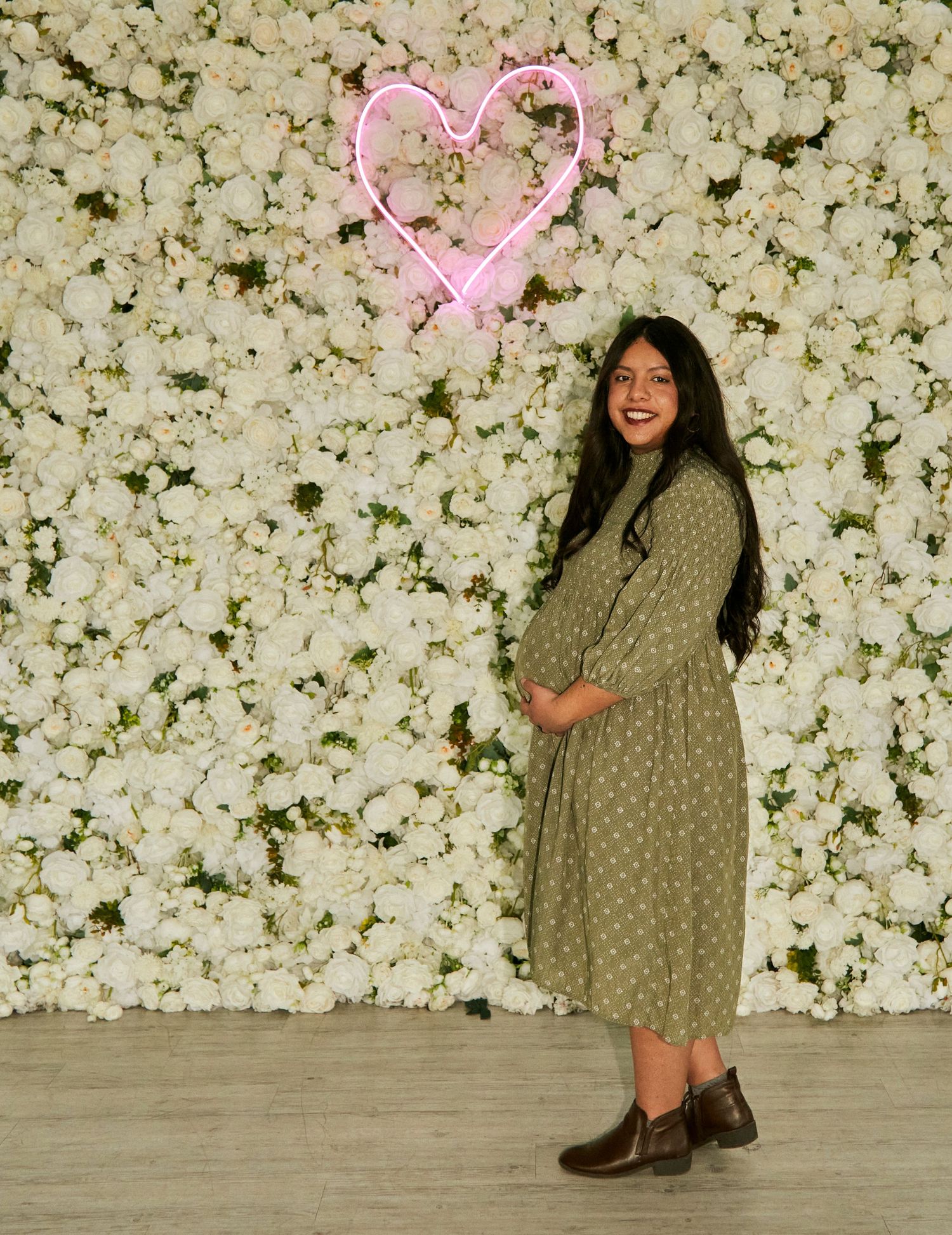 A maternity photo featuring a green dress against a white floral wall with pink neon heart.