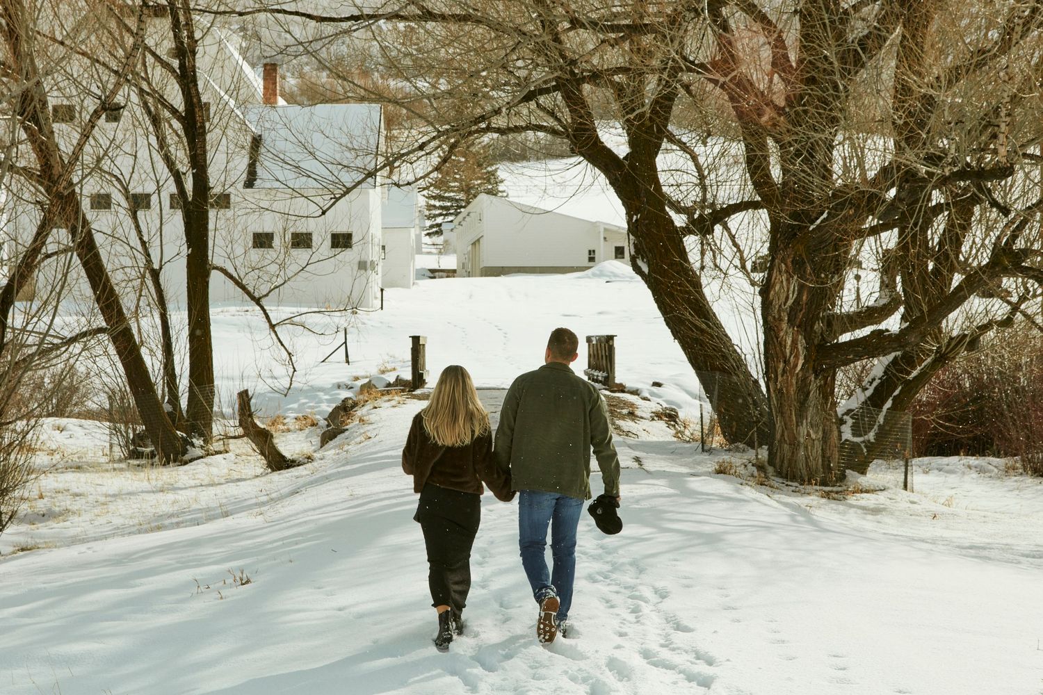 A couple walks hand in hand down a snowy path with white buildings and bare trees in the winter landscape.