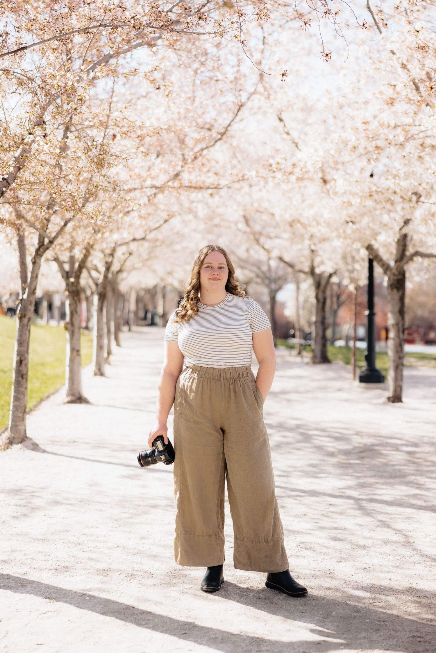 Photographer with camera stands on tree-lined path with blooming spring cherry blossoms overhead.