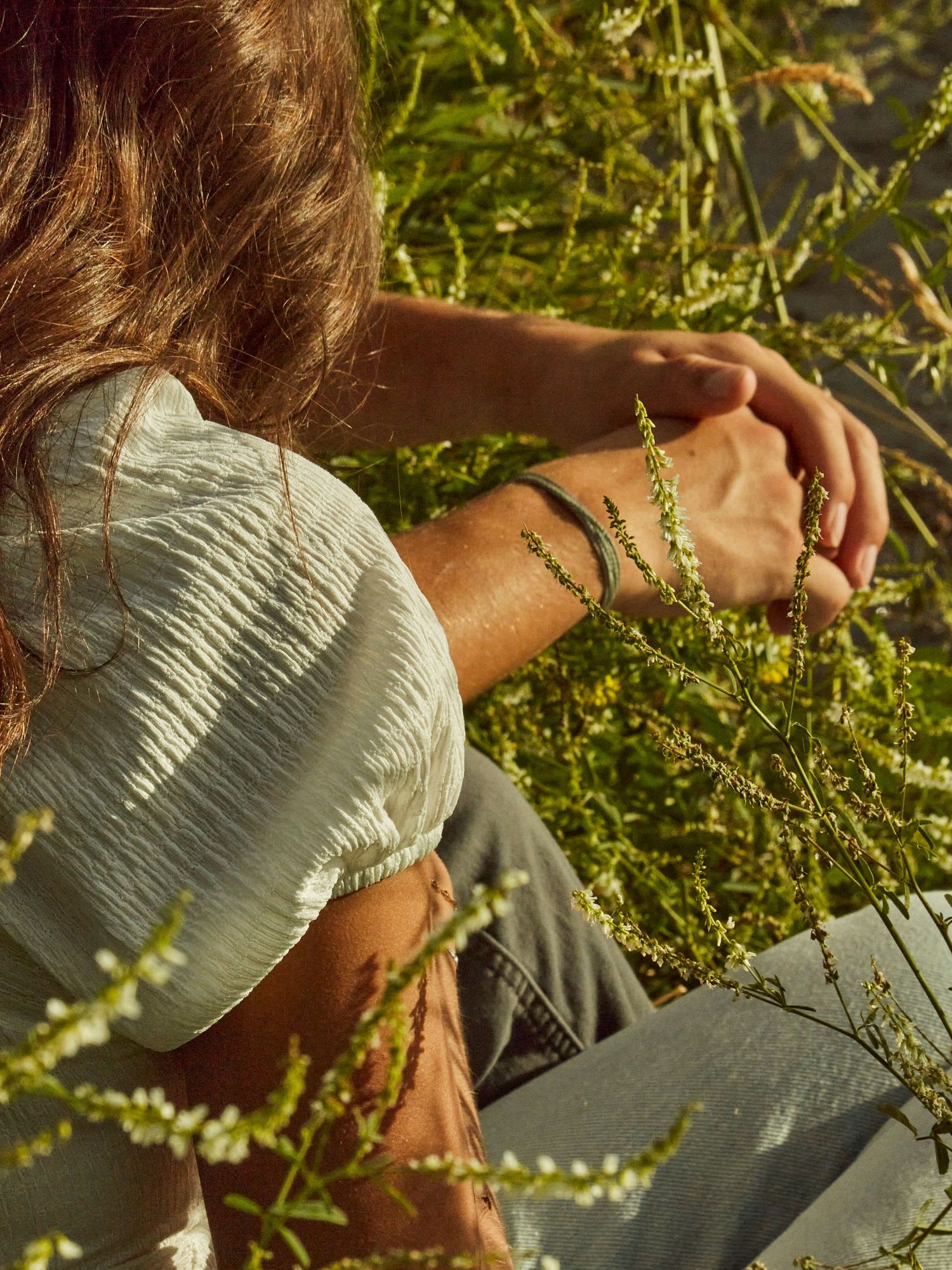 Person sitting in sunlit grass reaching toward delicate wildflowers.
